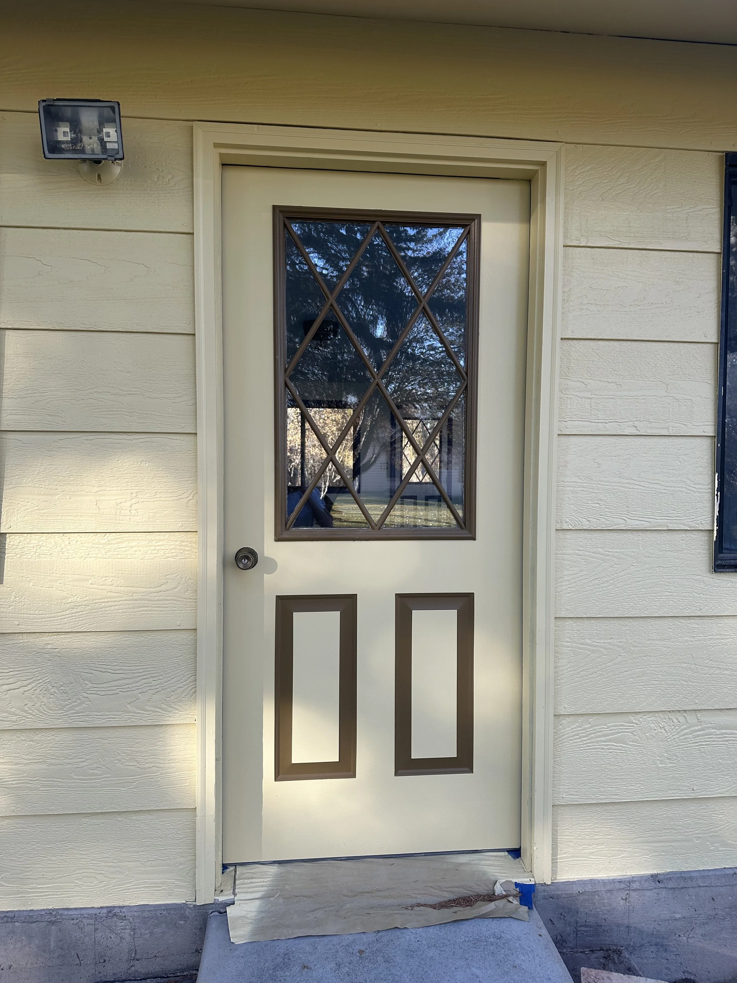 Exterior view of a house with a cream-colored door with a diamond-patterned window at the top and two rectangular panels below. The house has beige horizontal siding, and there is a security light mounted on the wall to the top left of the door. The 
