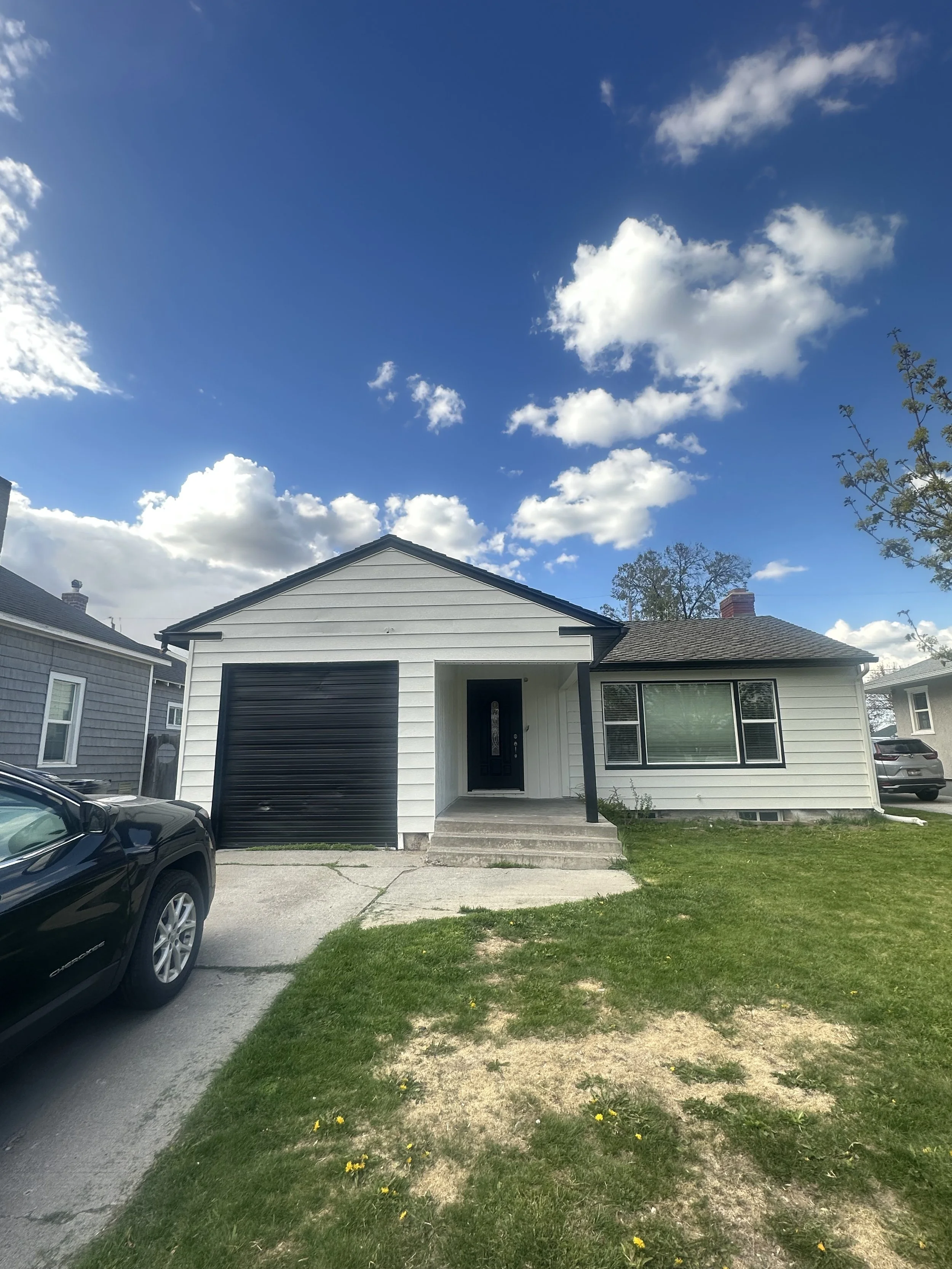 A house with white siding, a black garage door, and a black front door, with a driveway and a lawn with patches of dirt and some yellow flowers under a partly cloudy blue sky.
