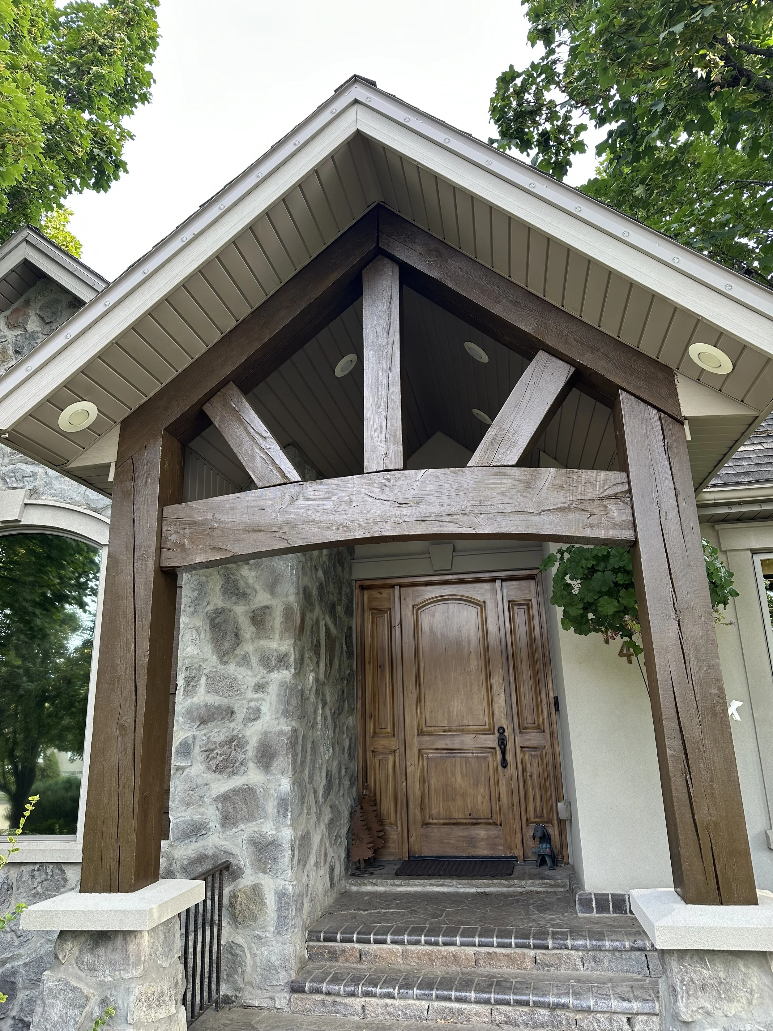 Front porch of a house with a stone wall, wooden front door, and a large wooden beam structure supporting the roof over the entrance.