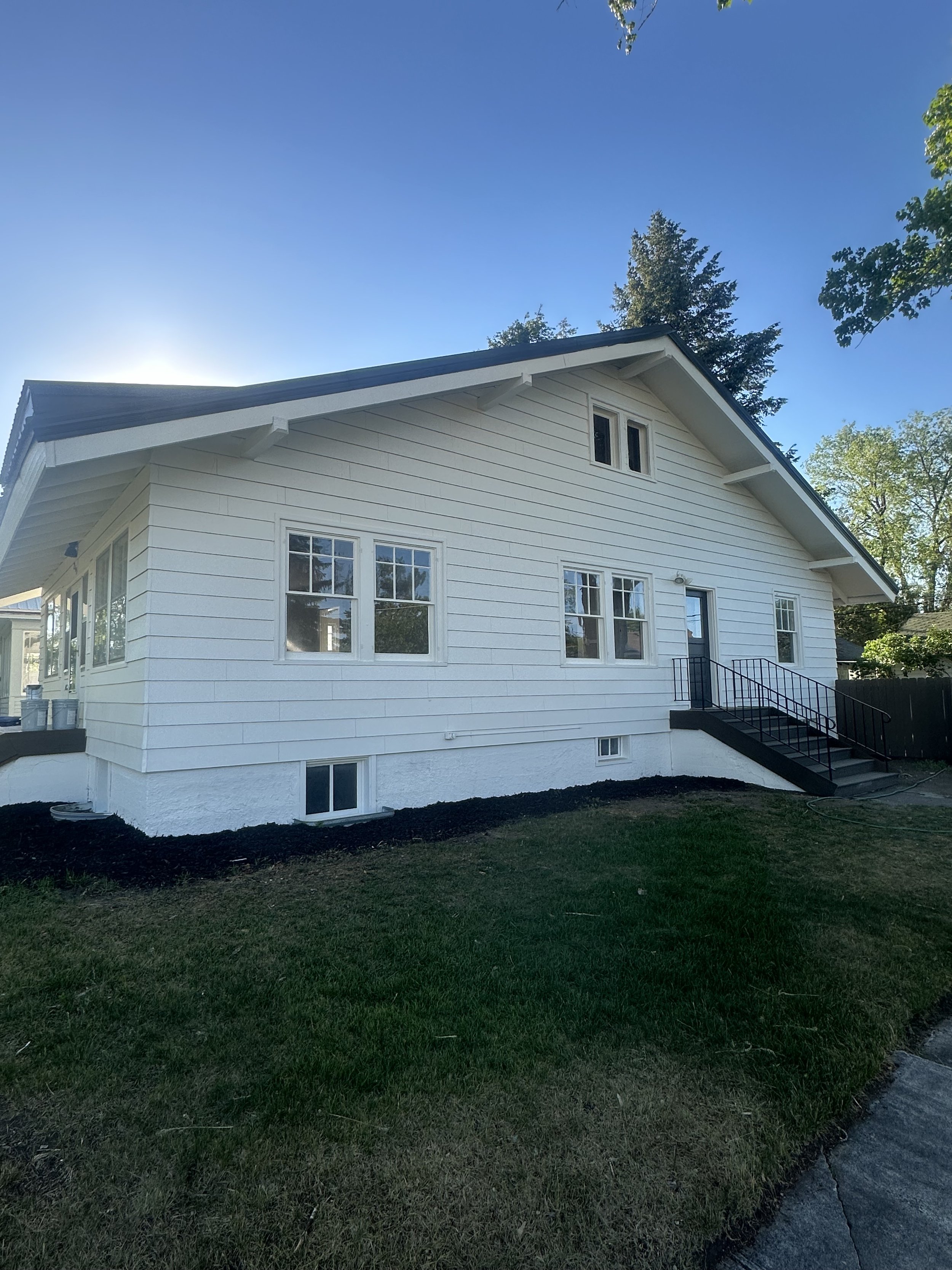 A white house with a sloped roof, multiple windows, and stairs leading to a door, with a grassy yard and trees in the background.