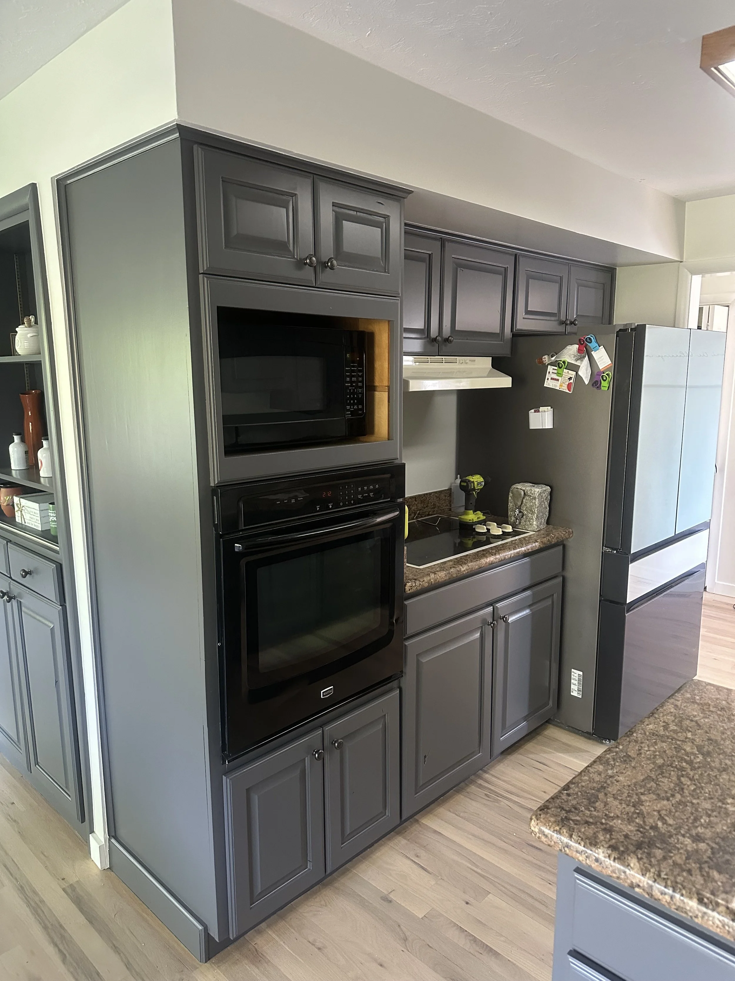 Gray kitchen cabinets with built-in black microwave and oven, granite countertops, and a side-by-side refrigerator. The kitchen has a wood floor and various small objects on the counter.