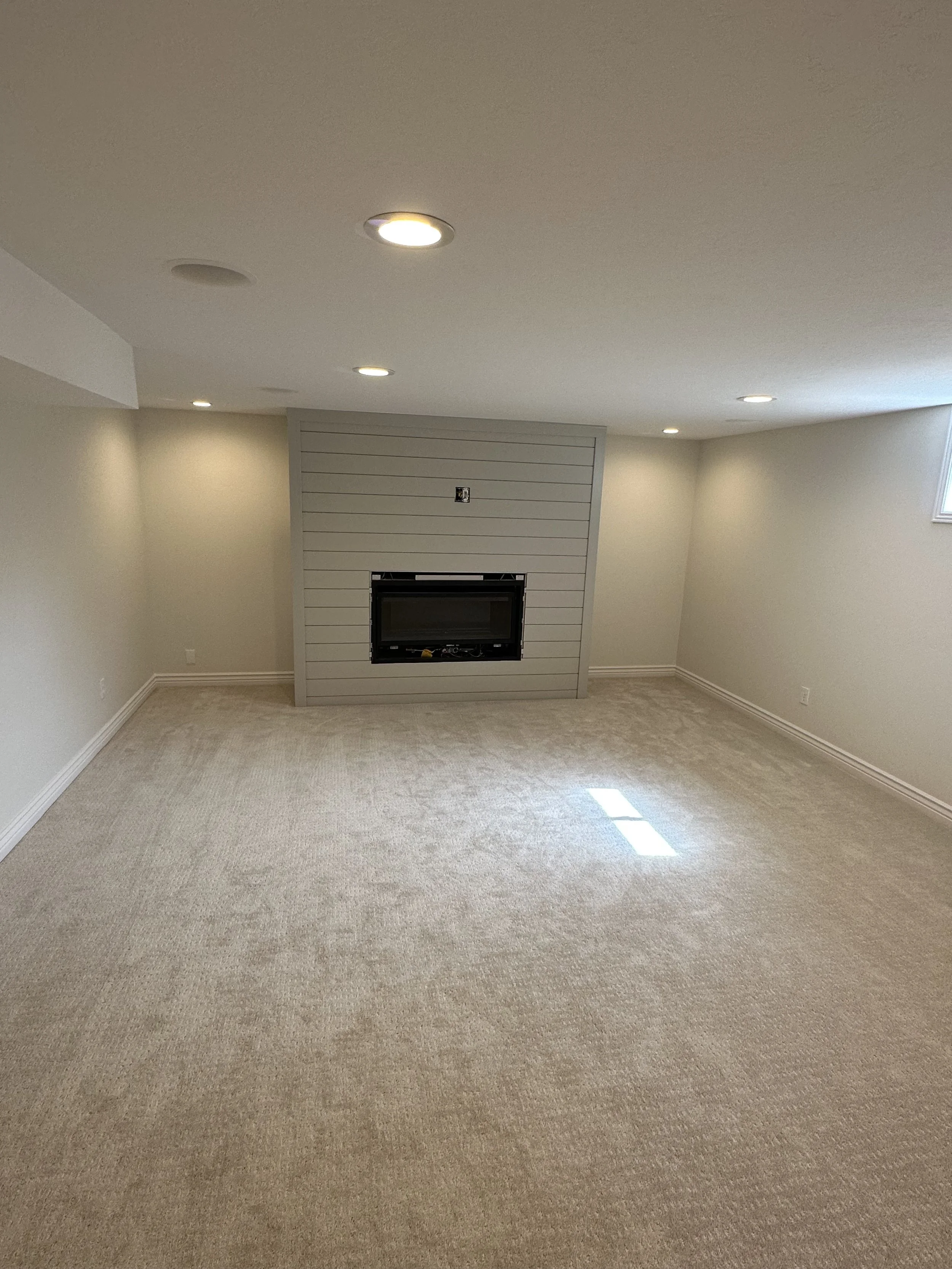 Empty basement room with beige carpet, white walls, a fireplace centered on a gray shiplap wall, and recessed lighting in the ceiling.