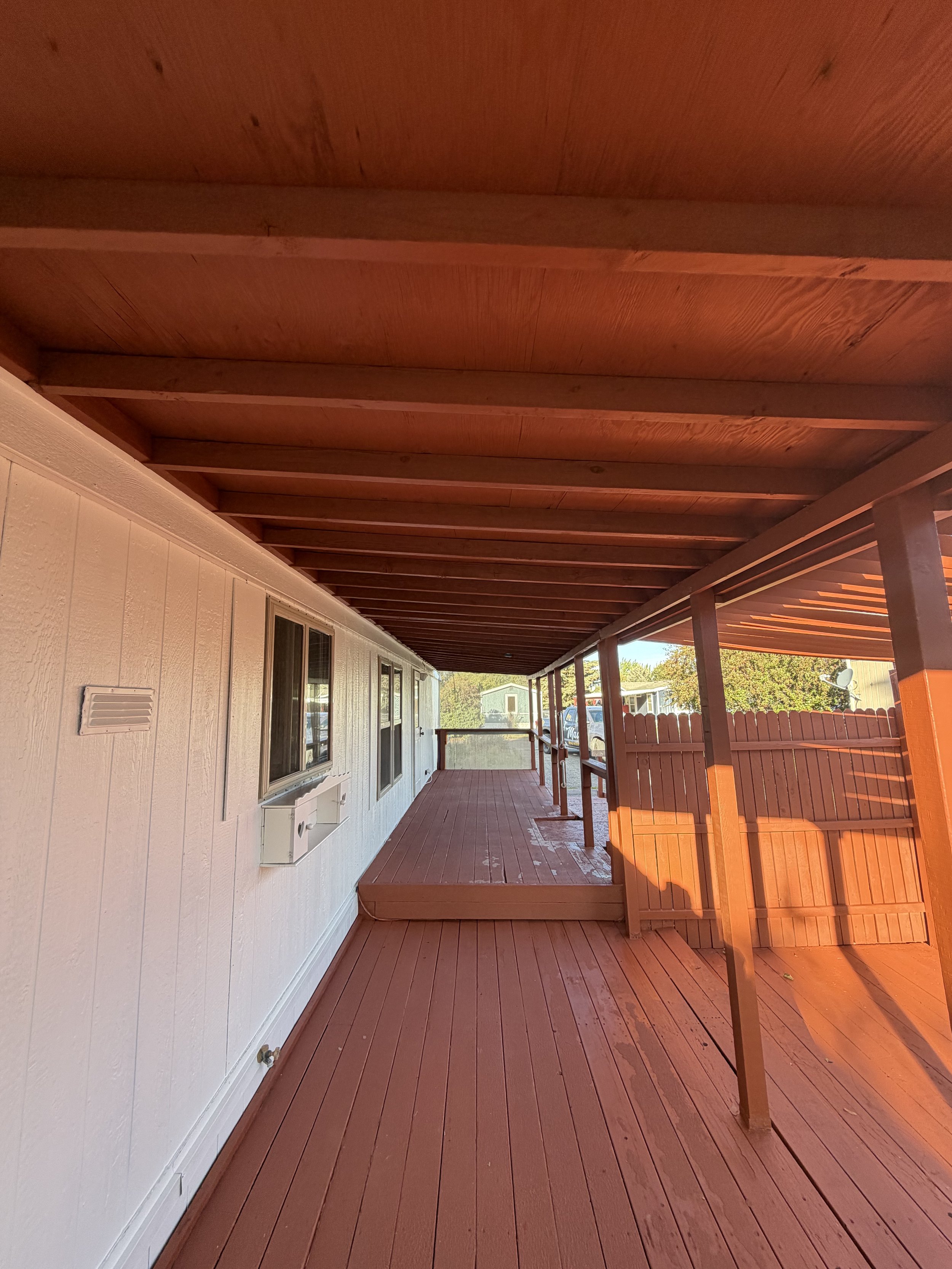 A wooden deck attached to a house with a railing and a small step, with a white wall featuring windows and a vent, and outdoor scenery including trees and neighboring houses in the distance.