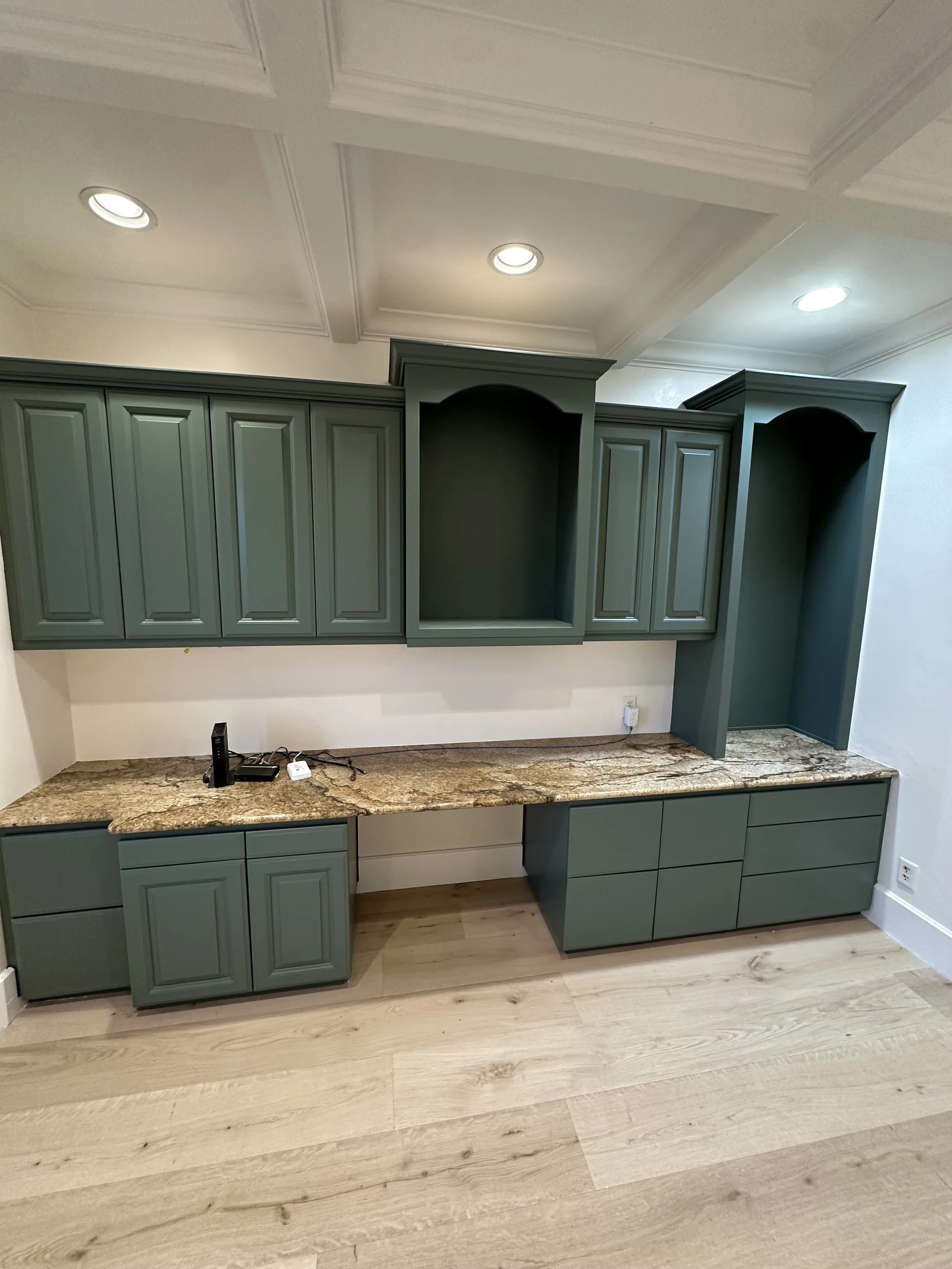 Green cabinetry with a granite countertop in a kitchen or office space, with recessed ceiling lights.