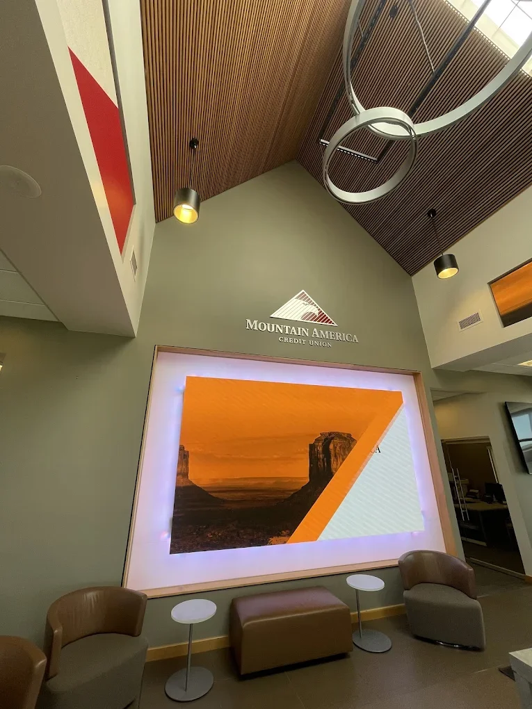 Interior lobby of a Mountain America Credit Union branch with a large digital screen showing a landscape, seating area with two chairs, a bench, and small round tables, wood-paneled ceiling, and modern lighting.