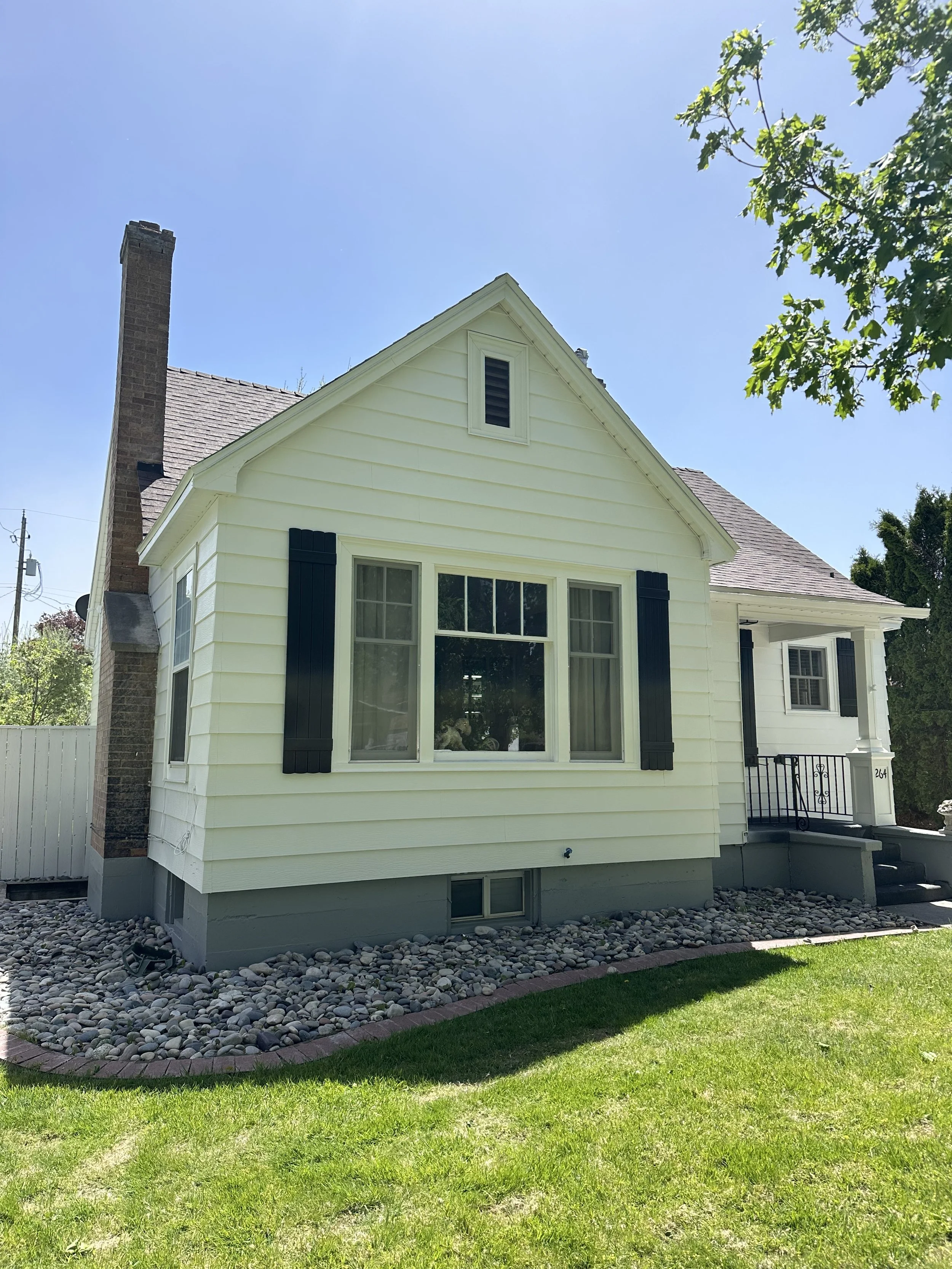 Front view of a white house with black shutters, a brick chimney, a small stairway, and a yard with green grass and rocks, under a clear blue sky.