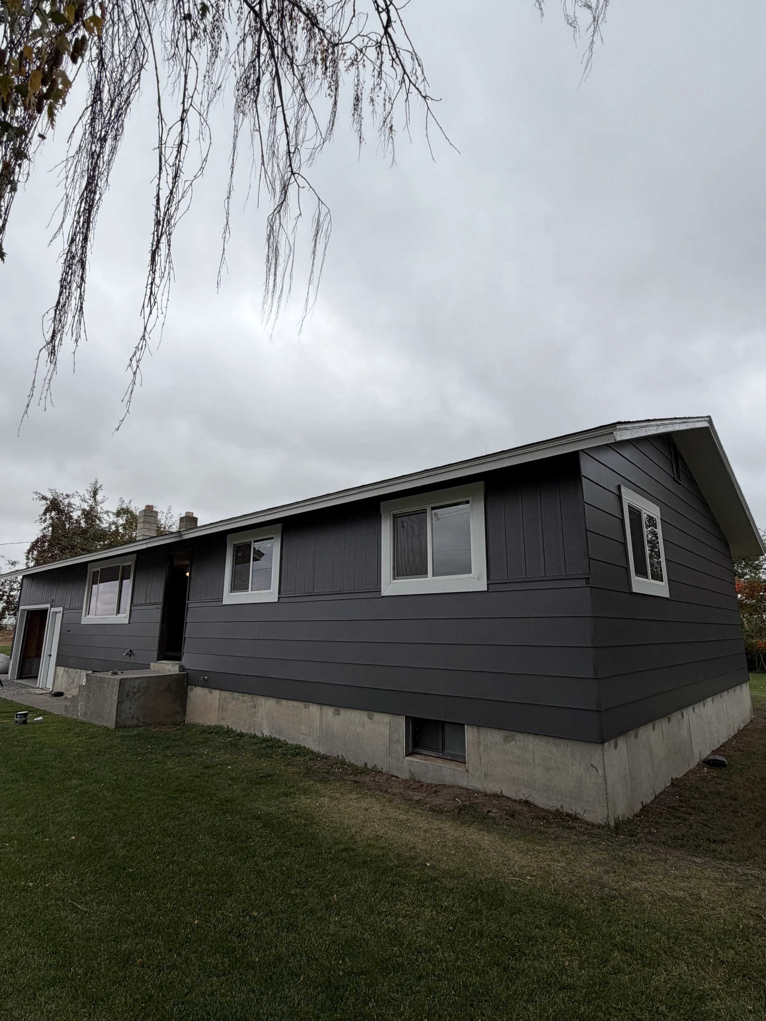 A dark gray, mid-century modern house with a sloped roof and white window frames, sitting on a concrete foundation with a grassy yard, under a cloudy sky.