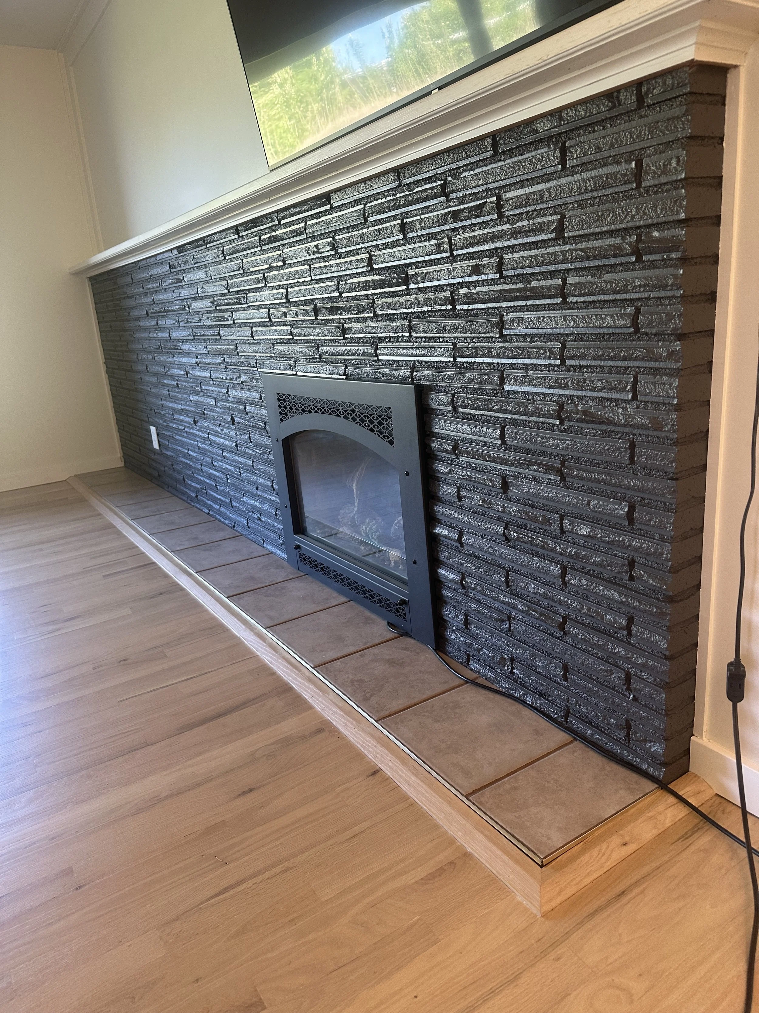 Photo of a black brick fireplace with a metal firebox insert, surrounded by a wooden hearth and beige flooring, with a television mounted above.