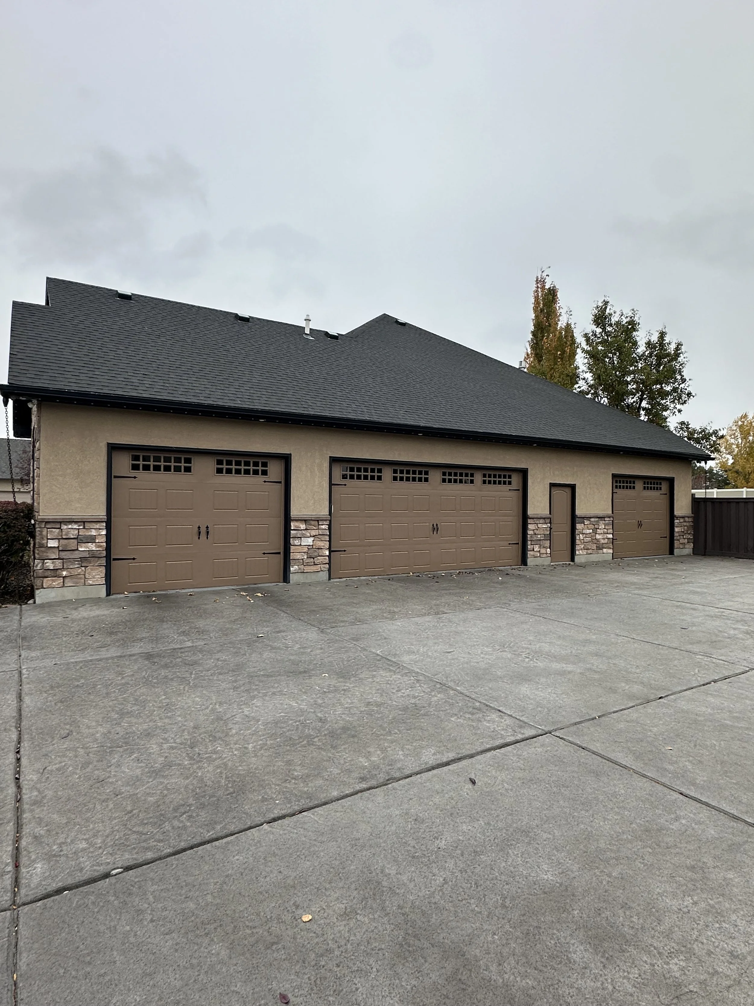 A large garage with three brown doors, a small door between the second and third doors, stone accents at the base, and a concrete driveway in front.