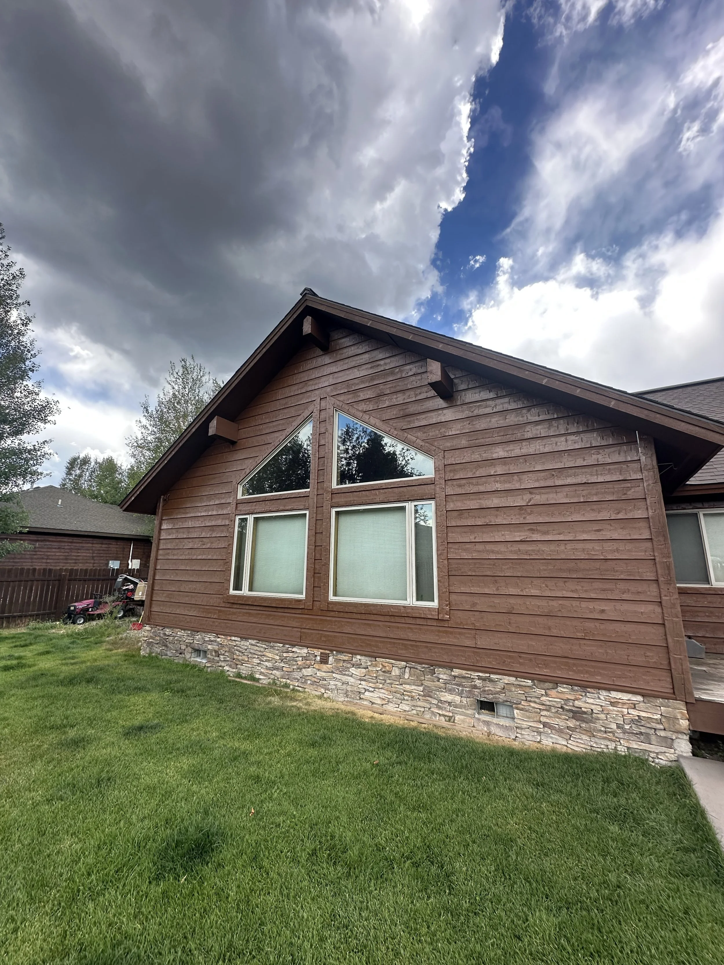 A house with brown wooden siding, stone foundation, and large windows, under a partly cloudy sky.