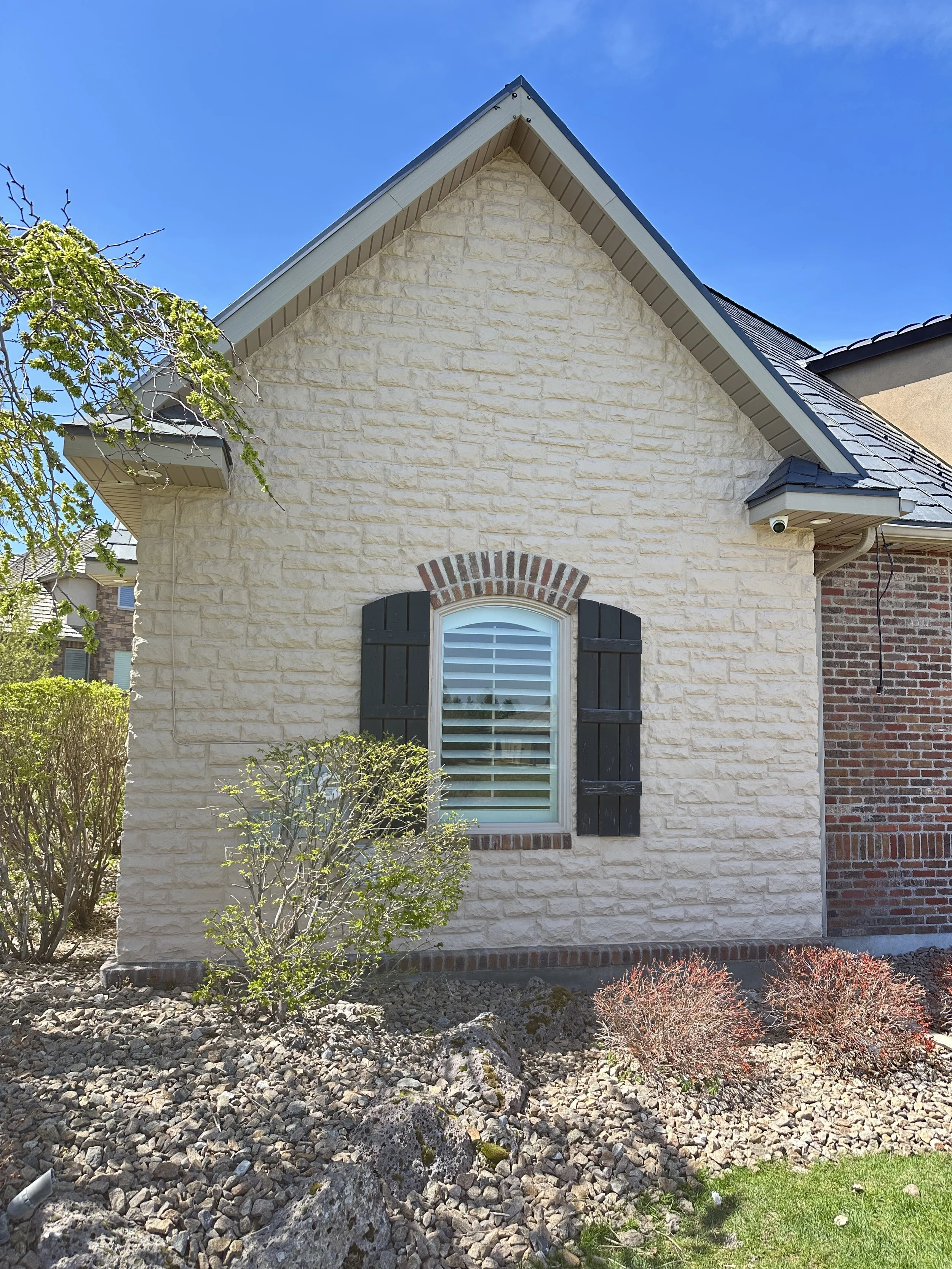 Front view of a house with cream brick exterior, black shutters, and a window with white blinds, surrounded by landscaped rocks and bushes under a clear blue sky.