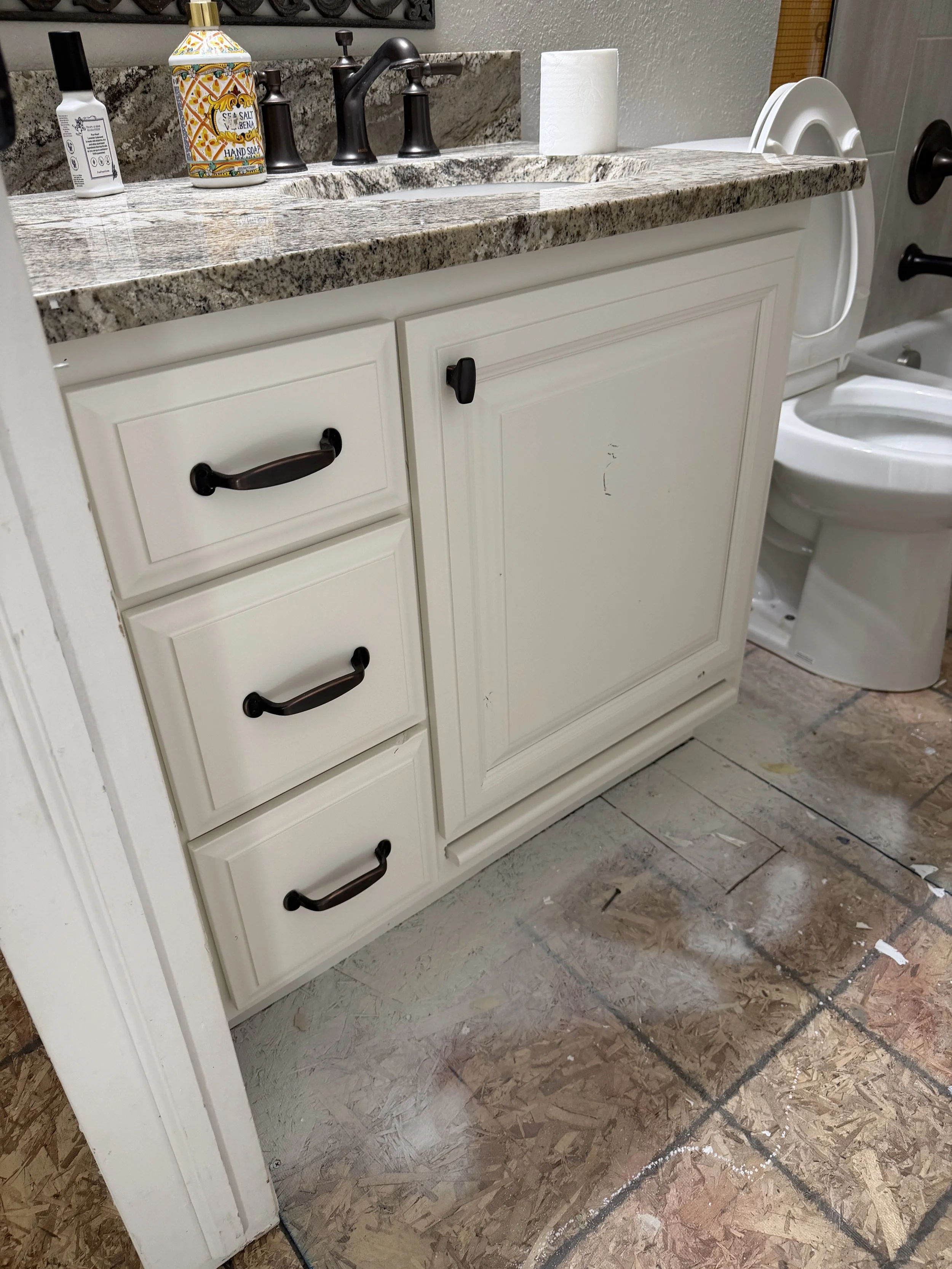 Bathroom vanity with a granite countertop, four drawers with black handles, and a cabinet with a black knob. Items on the counter include a hand soap dispenser, a decorative jar, a bottle of nail polish, a roll of paper towels, and a book. Part of a 