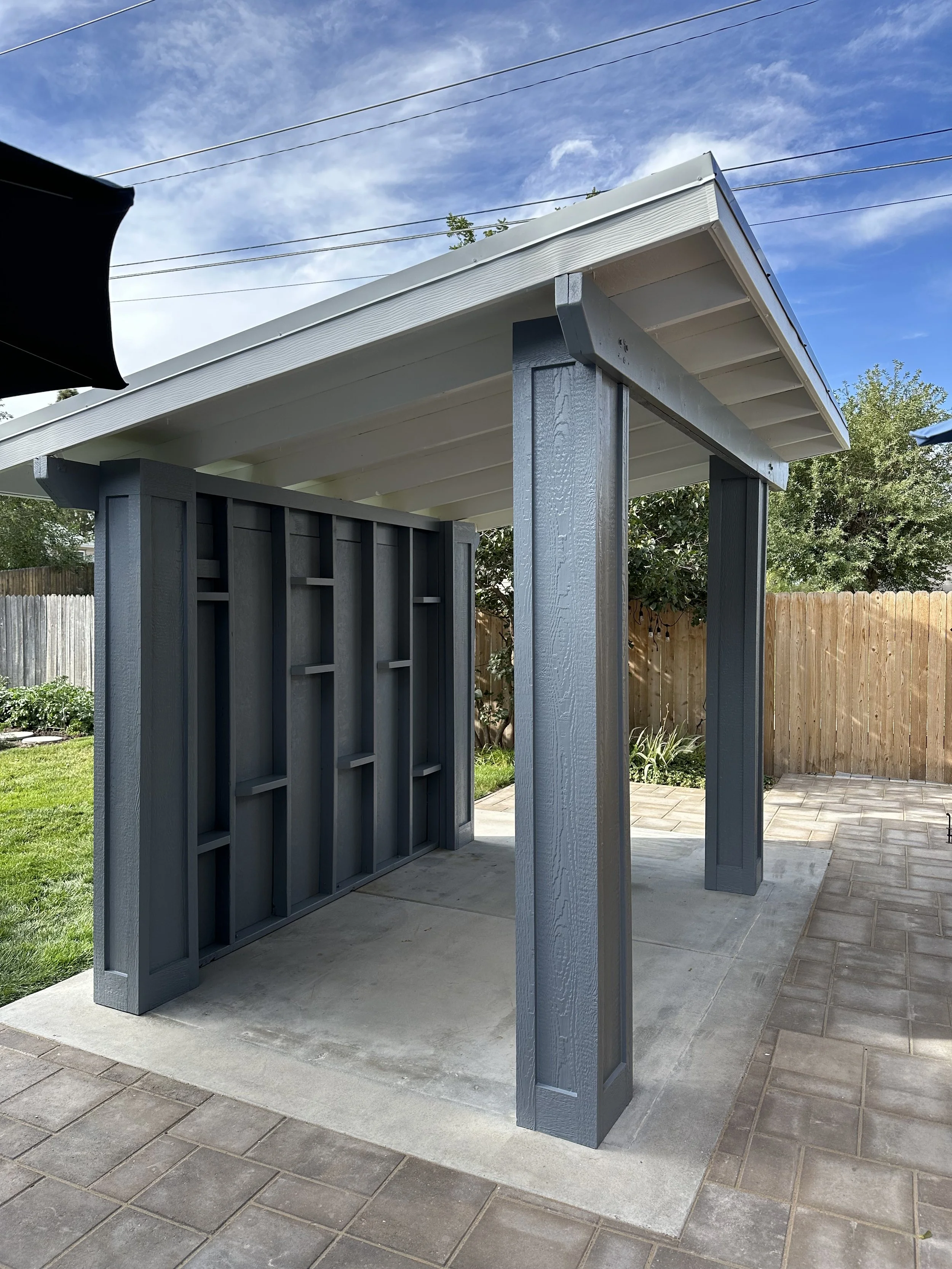 A small outdoor structure with gray wooden support beams, a white sloped roof, and a gray lattice wall beside it, situated on a concrete slab with a brick-paved area surrounding it, in a backyard with green grass, trees, and a wooden fence.