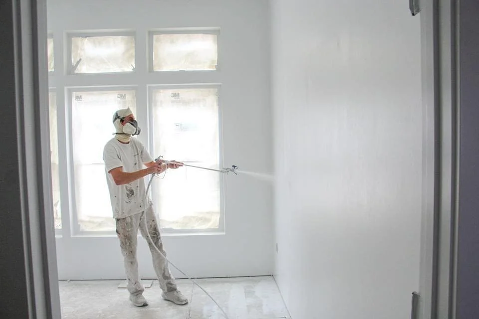 A person wearing protective gear, including a face mask and goggles, is using a spray gun to paint a white wall inside a room with large windows.