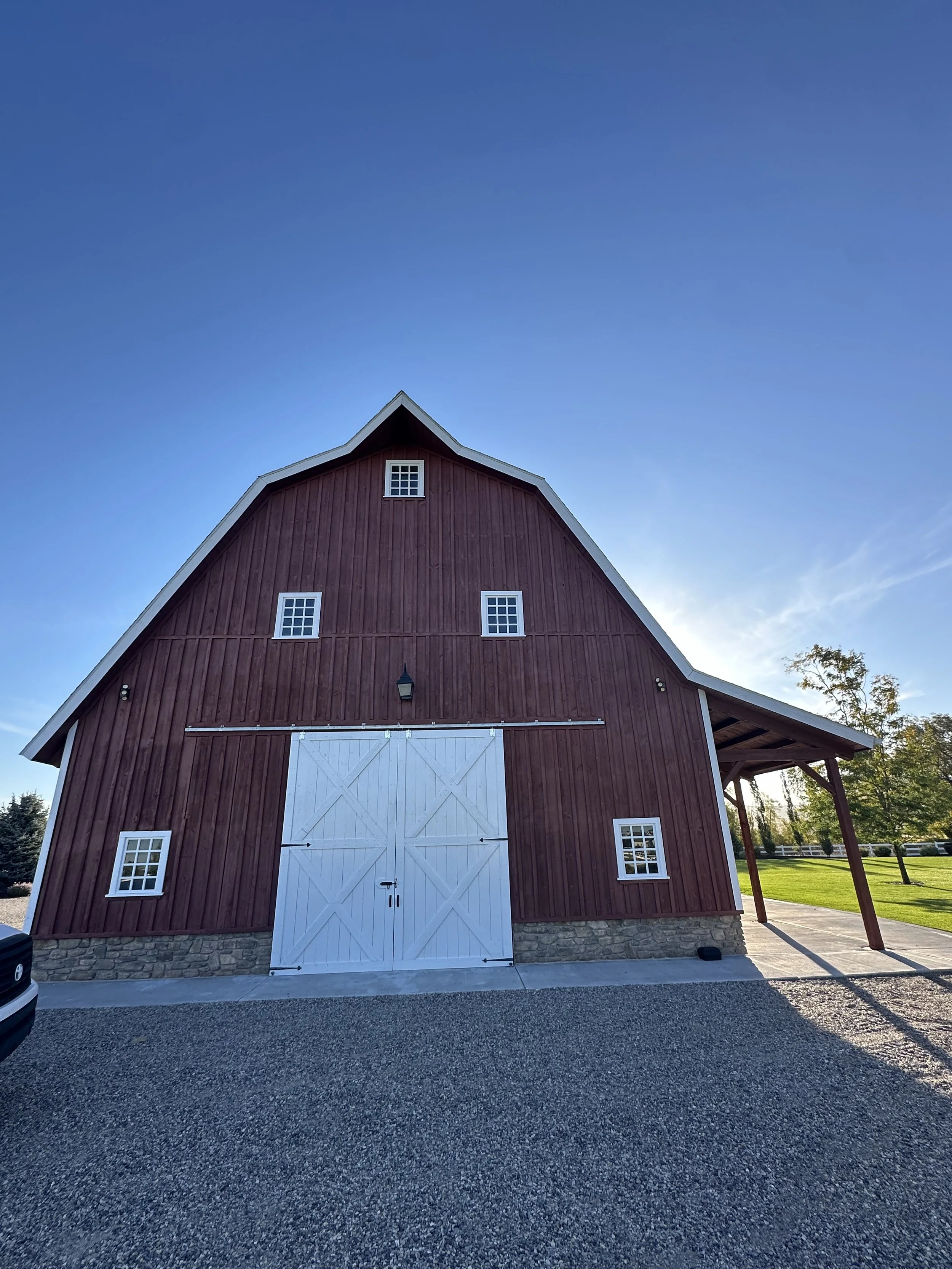 A red barn with white doors and windows, stone foundation, and a small porch with support beams, set against a clear blue sky and green trees in the background.