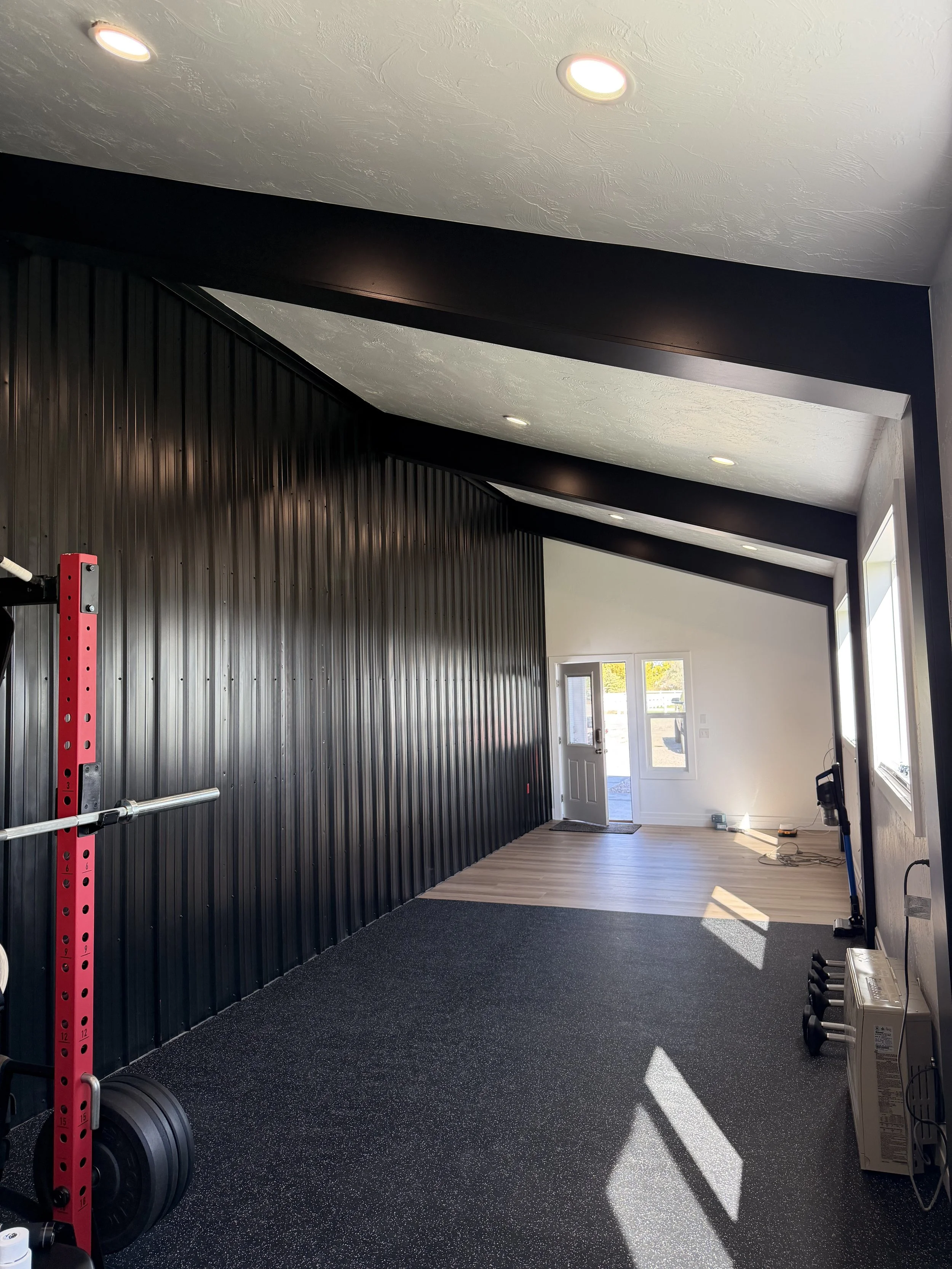 Empty gym with black wall panels, wooden flooring near the entrance, gym equipment, black workout mats, and sunlight streaming through the windows.