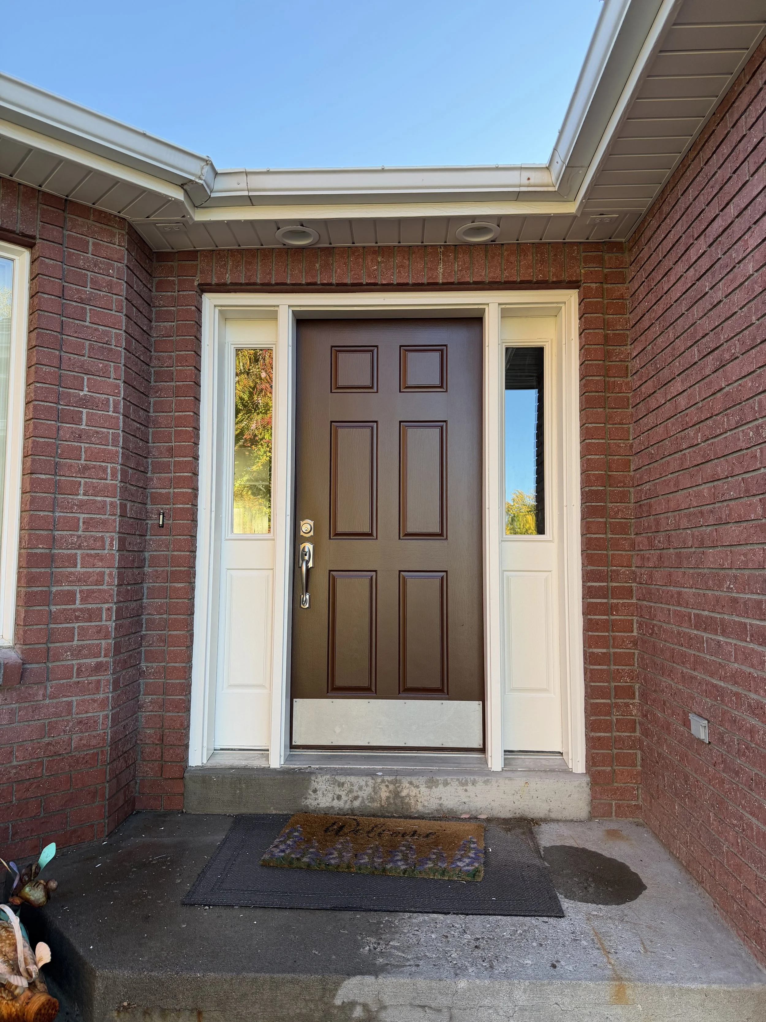 Front door of a house with a brown door surrounded by brick walls, a welcome mat on the concrete porch, and a blue sky overhead.