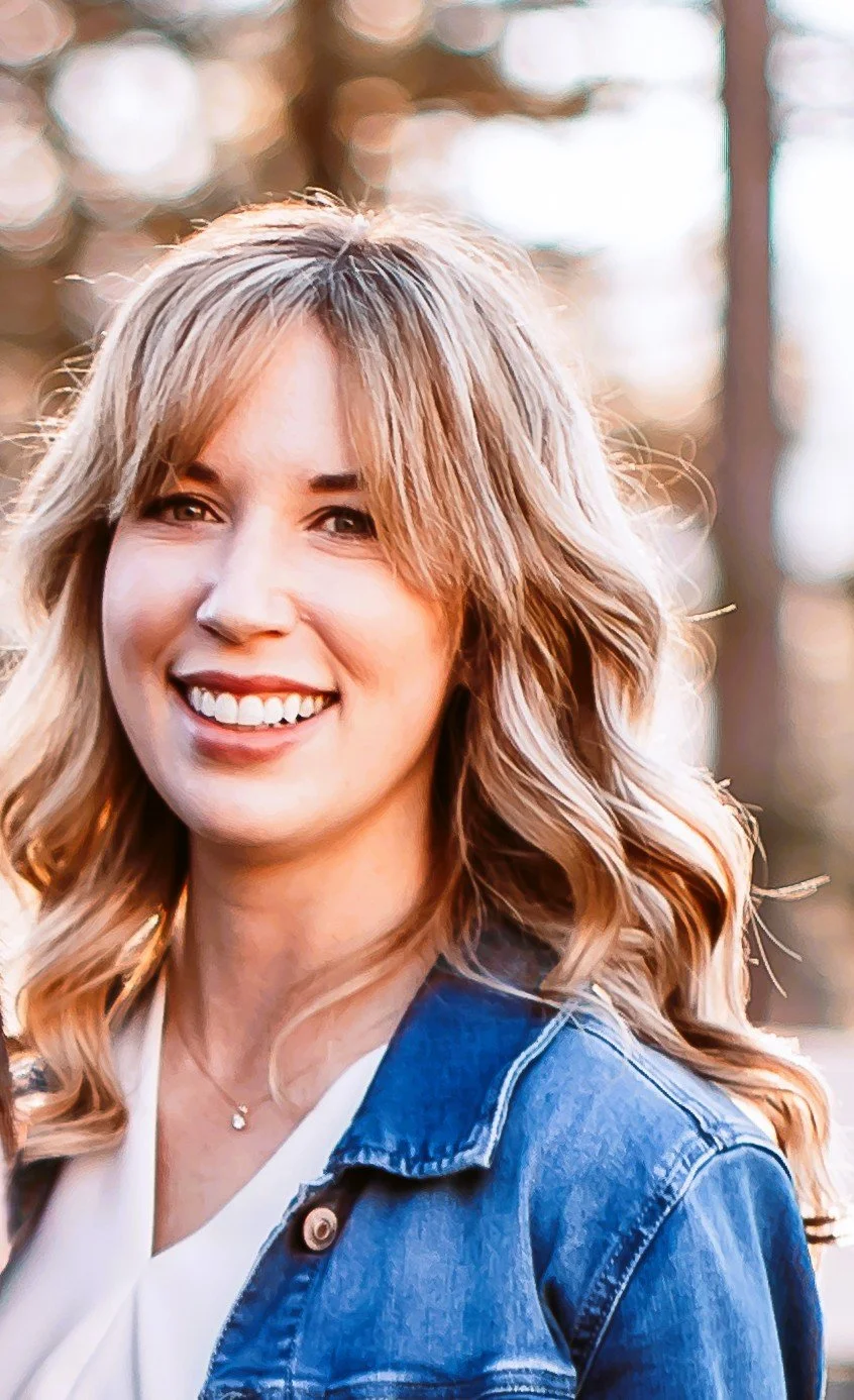 A woman with shoulder-length wavy blonde hair smiling outdoors, wearing a denim jacket and a white top.