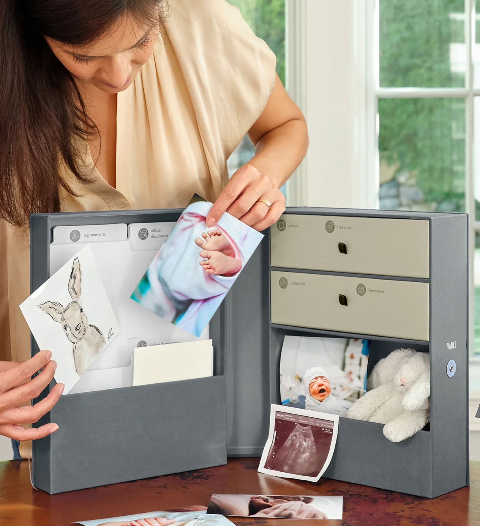 A woman organizing a memory box with baby photos, ultrasound images, and drawings on a table near a window.