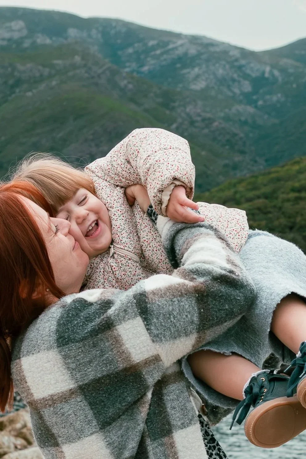 Une femme avec des cheveux rouges tient un jeune garçon dans ses bras près d'un lac entouré de montagnes.