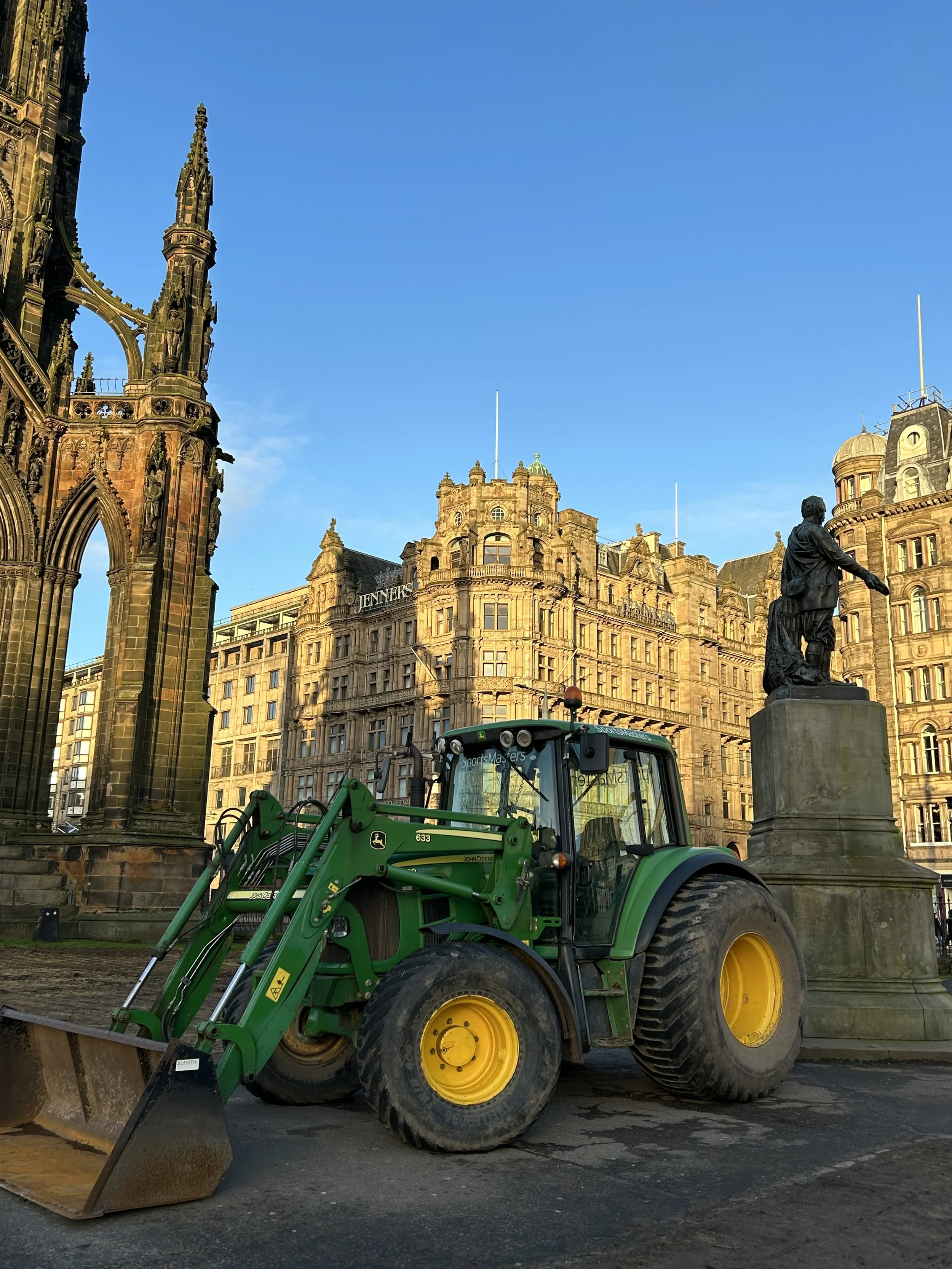 A green John Deere front loader parked on a city street near historic buildings and a statue.