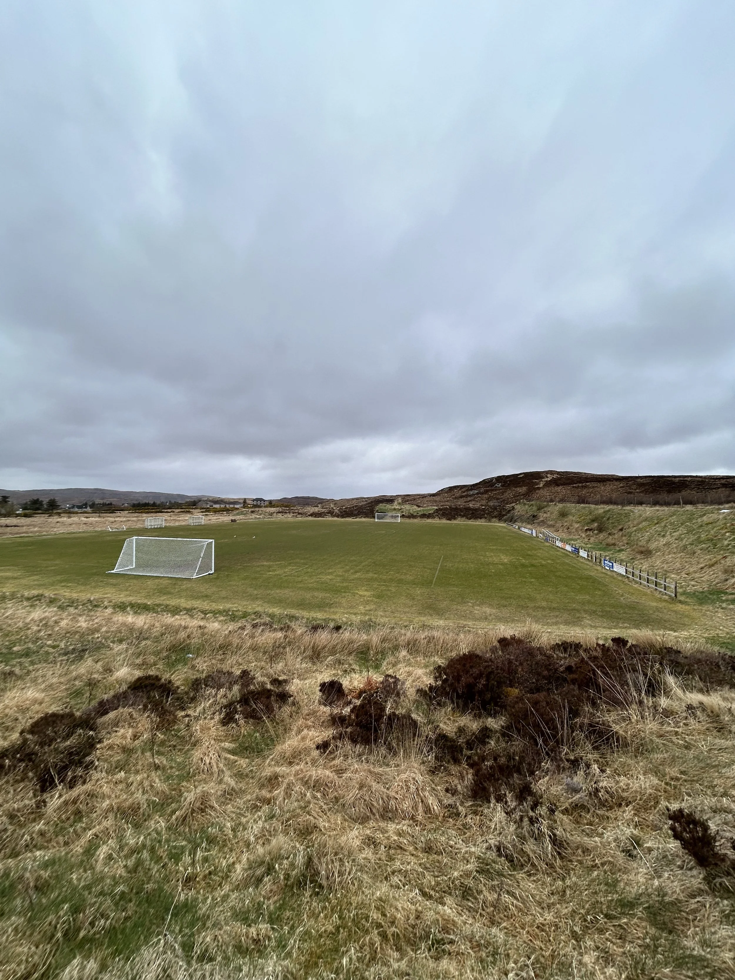 Open grassy field with soccer goals under a cloudy sky and some hills in the background.