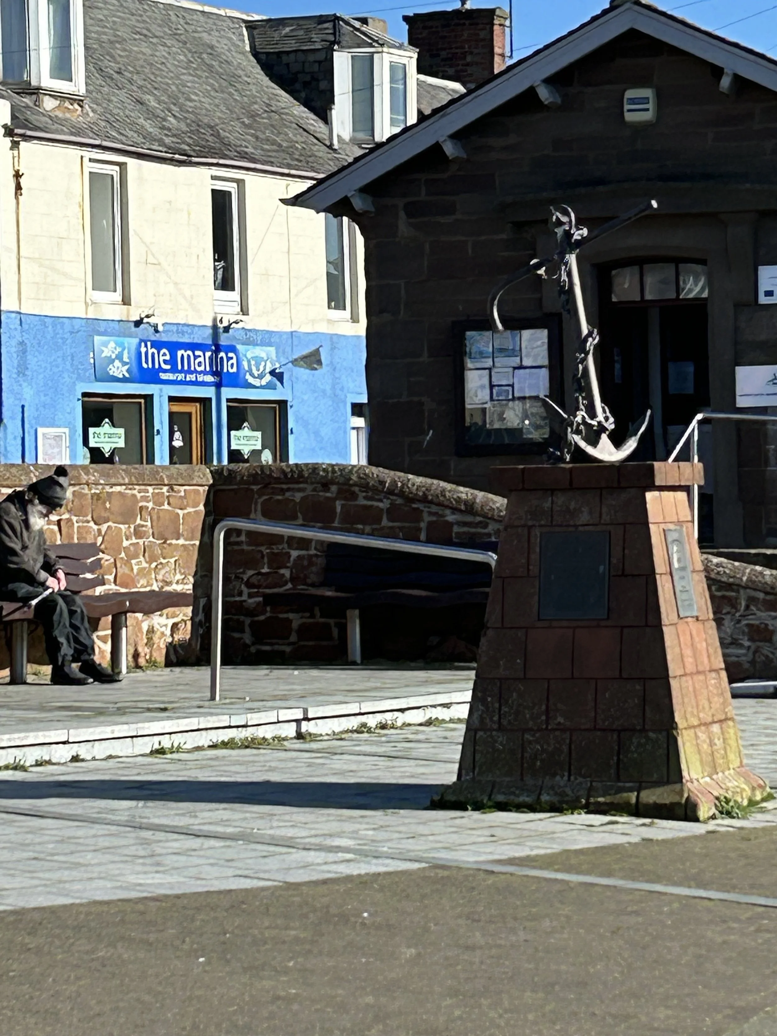 A small harbor with a modern anchor sculpture on a brick pedestal, a person sitting on a bench, and a building with a sign reading 'the marina' in the background.