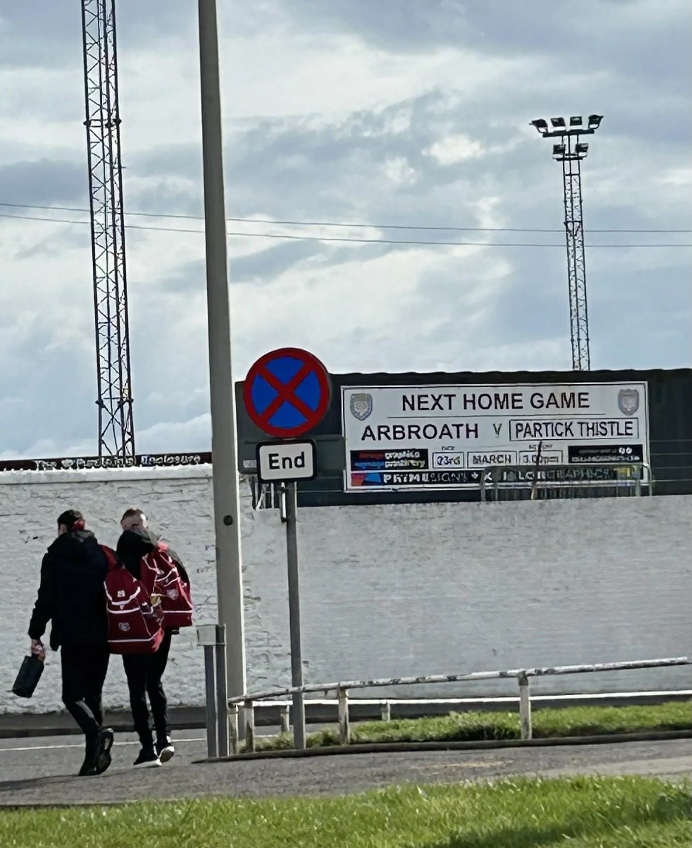 People walking near a signboard advertising a football match between Arbroath and Partick Thistle, with floodlights and cloudy sky in the background.