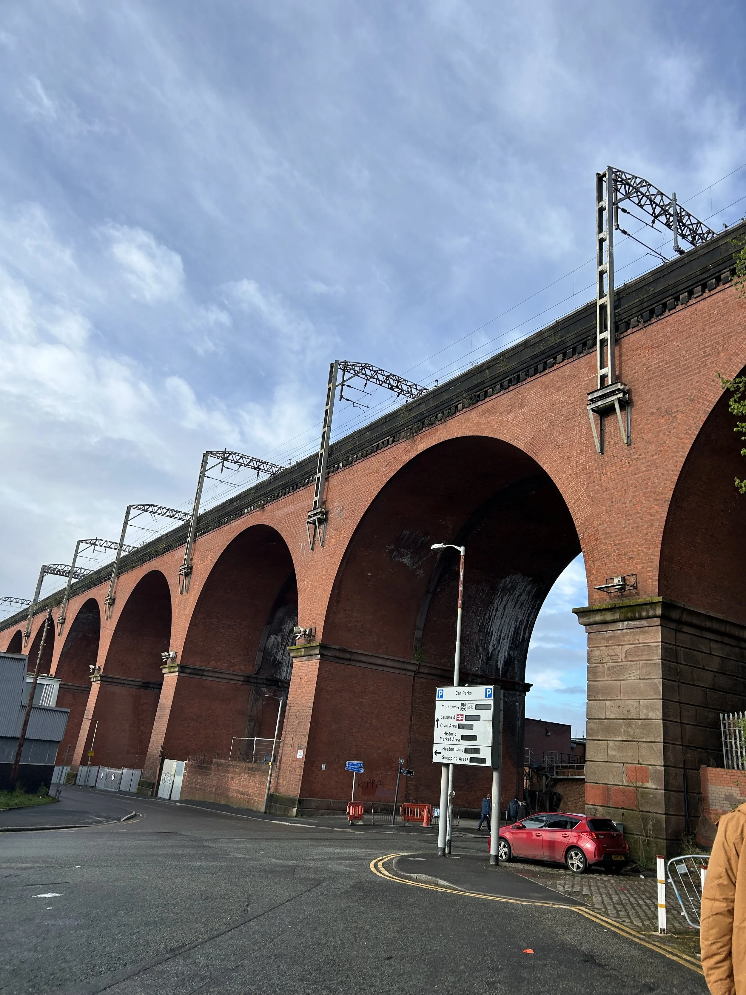 A large red brick viaduct with arches supports railway tracks above a street with cars and pedestrians, under a partly cloudy sky.