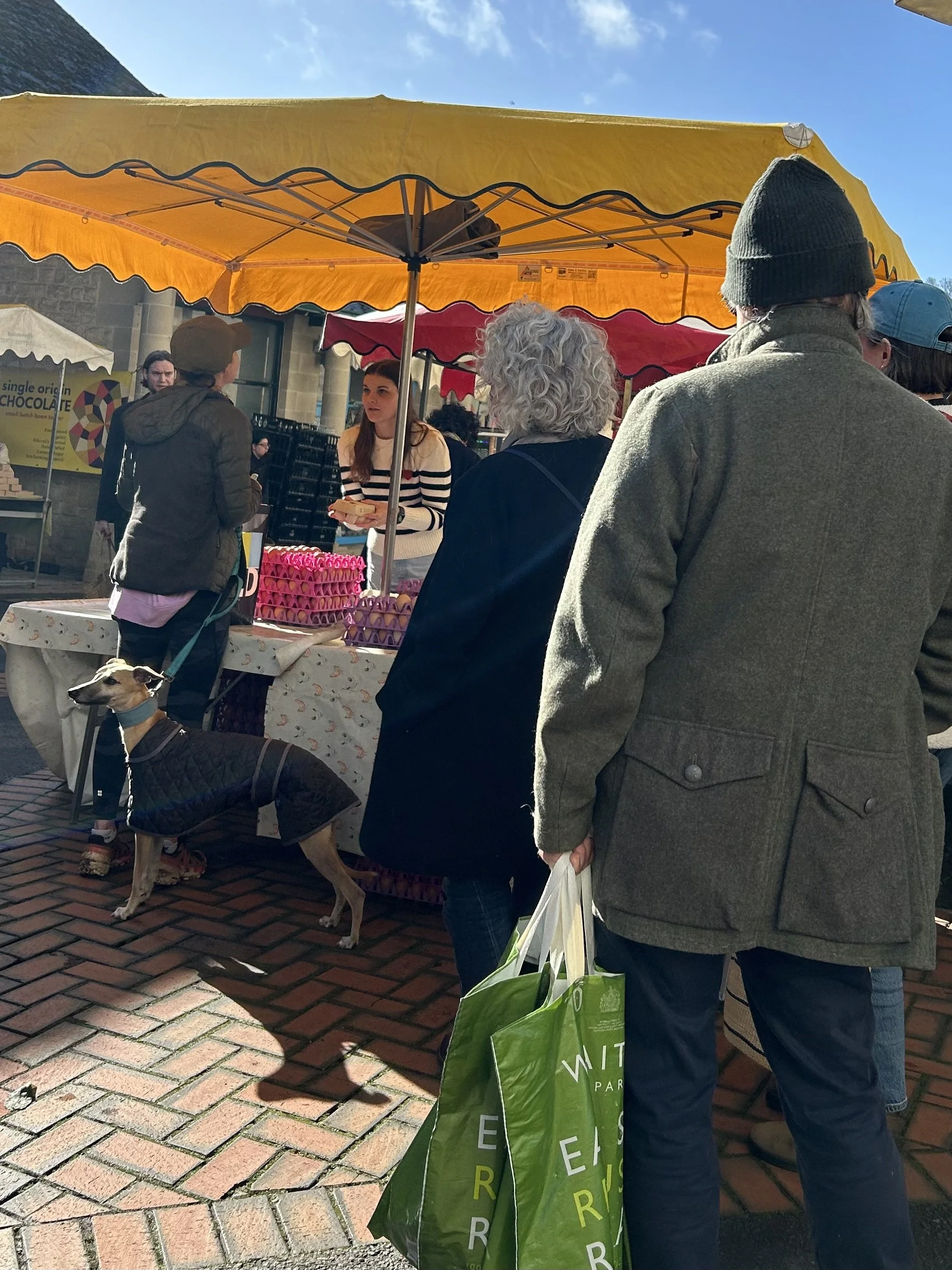 People shopping at an outdoor market stall under a yellow canopy, with a dog in a coat in the foreground and other shoppers carrying bags.