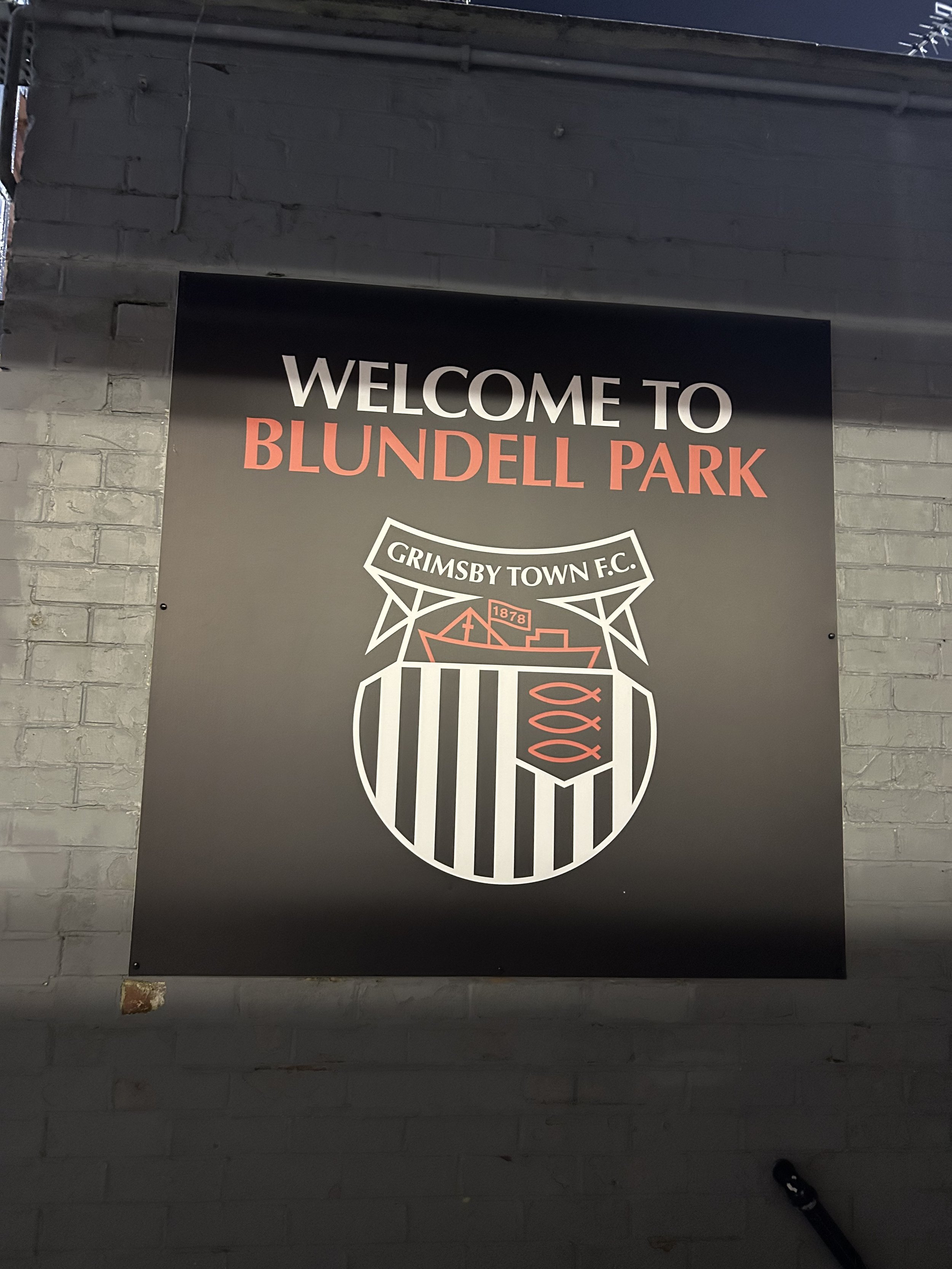 A black sign with white and red text welcoming visitors to Blundell Park, featuring the Grimsby Town F.C. emblem.