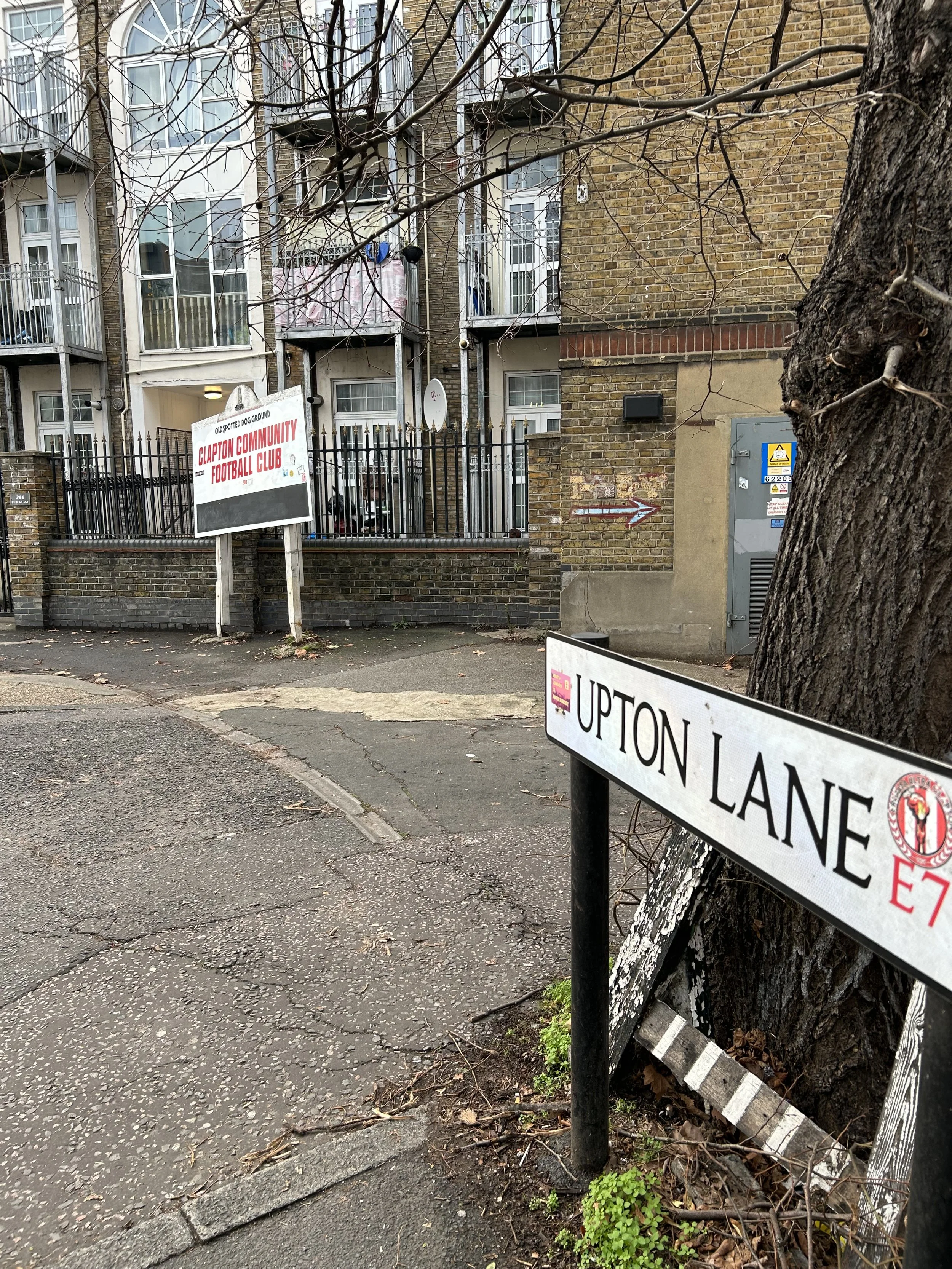 Street view showing a street sign for Upton Lane, a tree, a large building with apartment balconies and a sign indicating the location of Clapton Community Football Club in London.