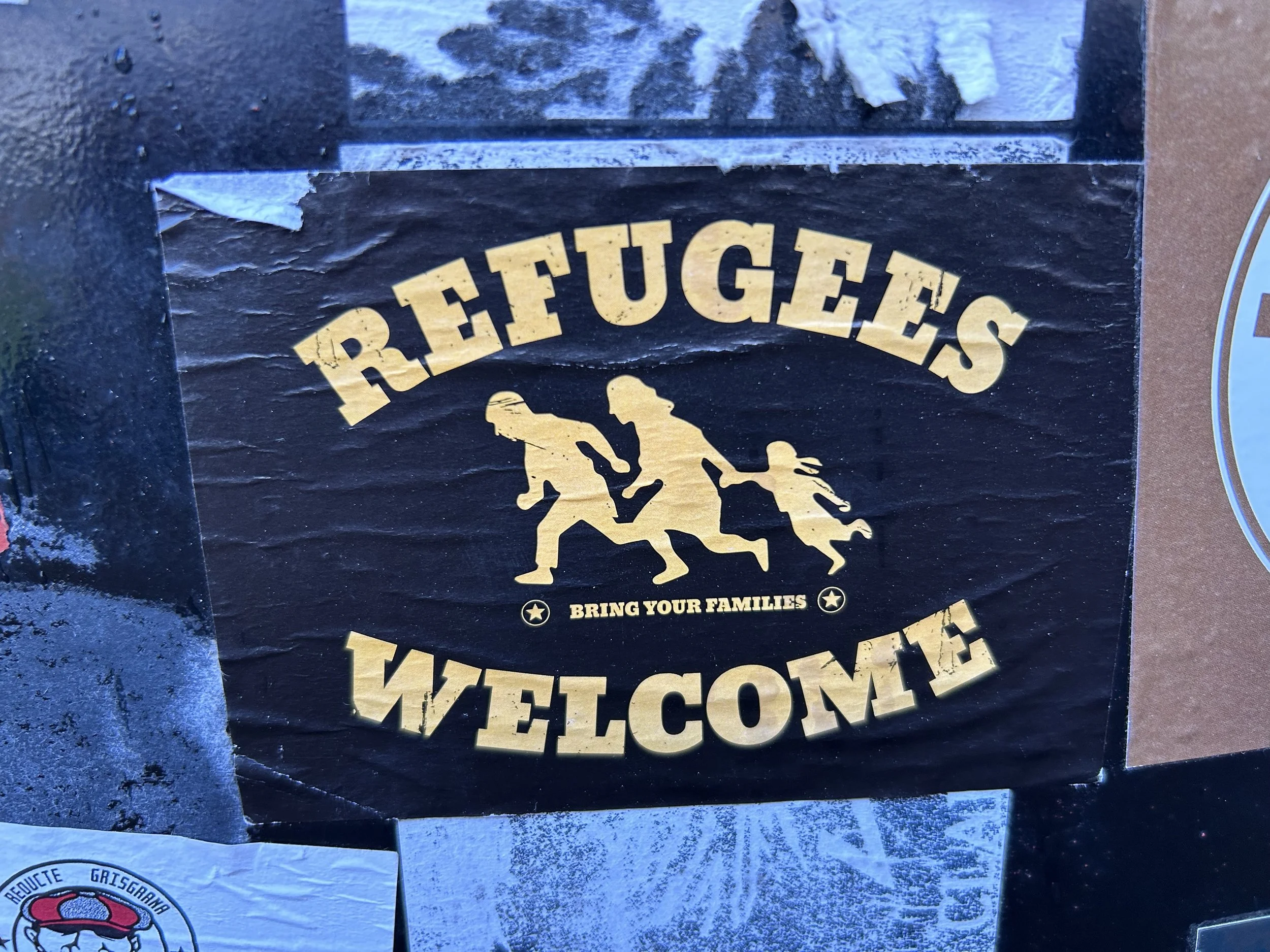 A printed sign with the words 'Refugees Welcome' in bold yellow letters, featuring a graphic of a refugee family holding hands, with the phrase 'Bring Your Families' below.