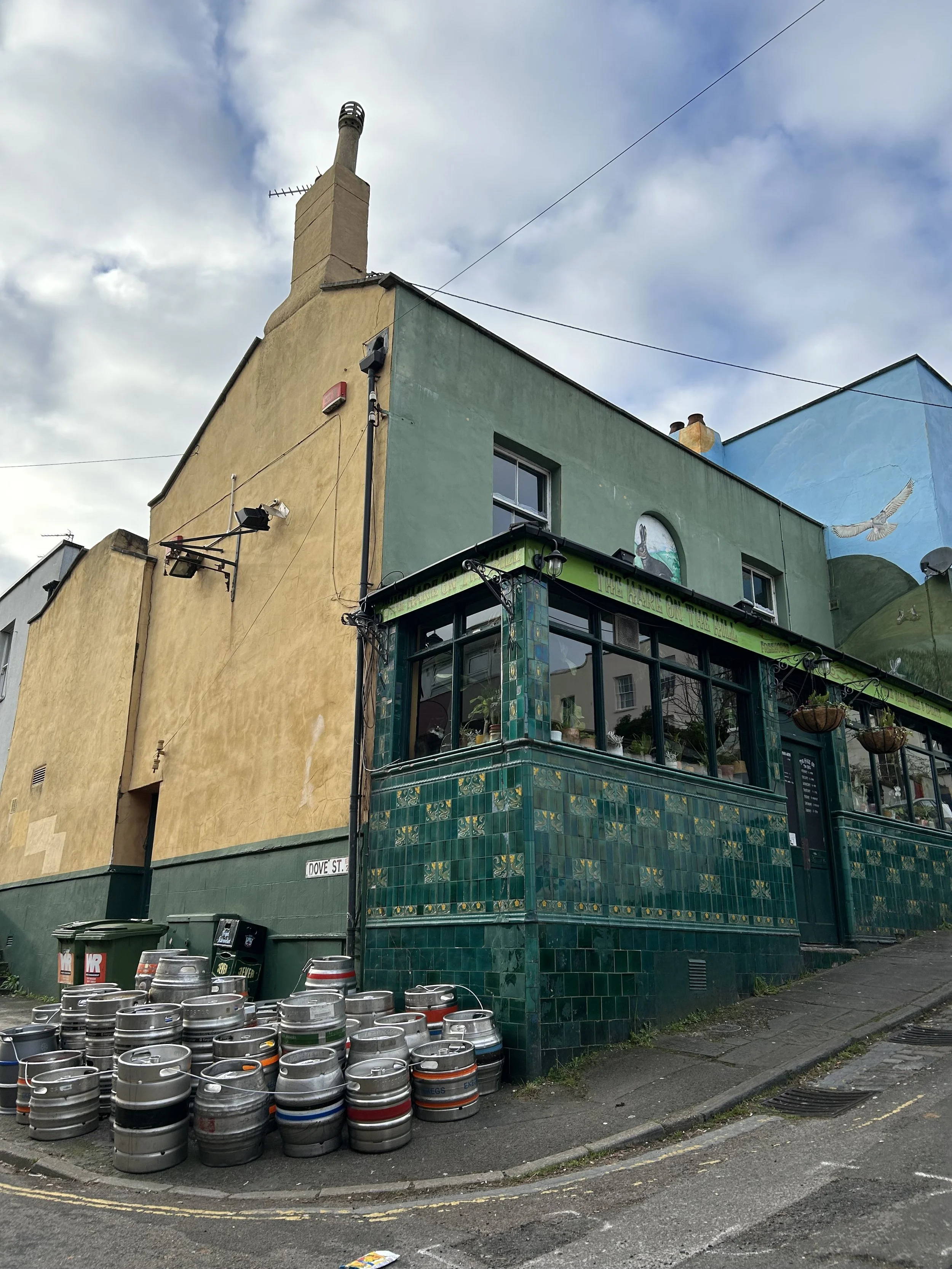 Colorful building with green tiles and glass enclosure on corner of street, with stacked beer kegs outside and murals on upper walls, cloudy sky above.