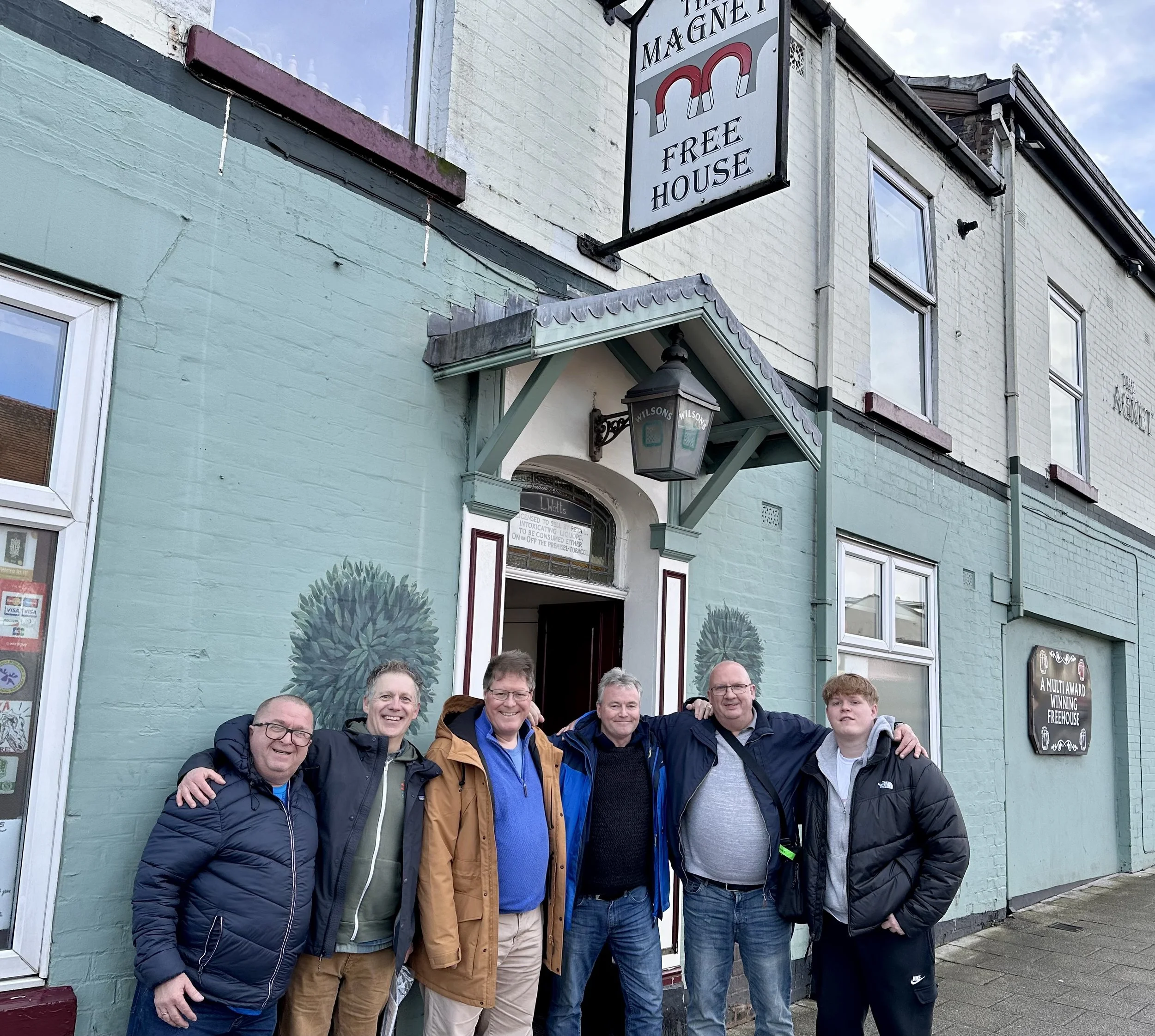 Group of six men standing outside a building labeled 'The Magnet Free House' with arms around each other's shoulders, smiling. The building has a mint green and white exterior with window art and a hanging lantern.