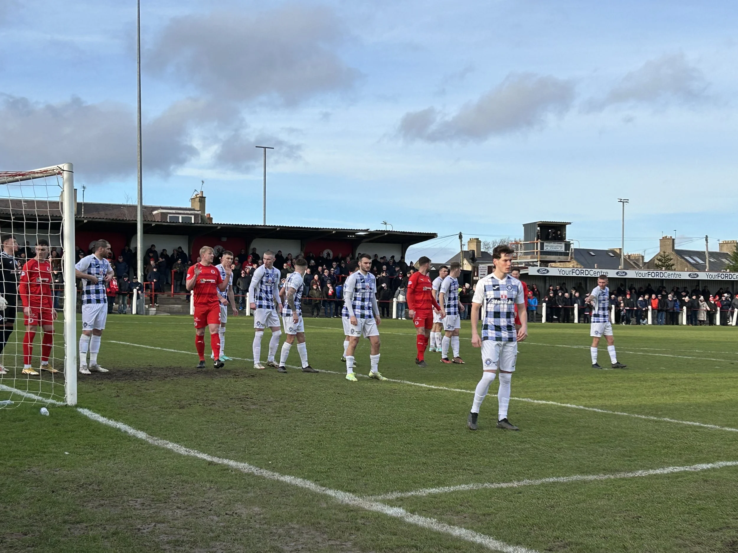 Soccer players on a field near the goal, with spectators in the stands watching in the background.