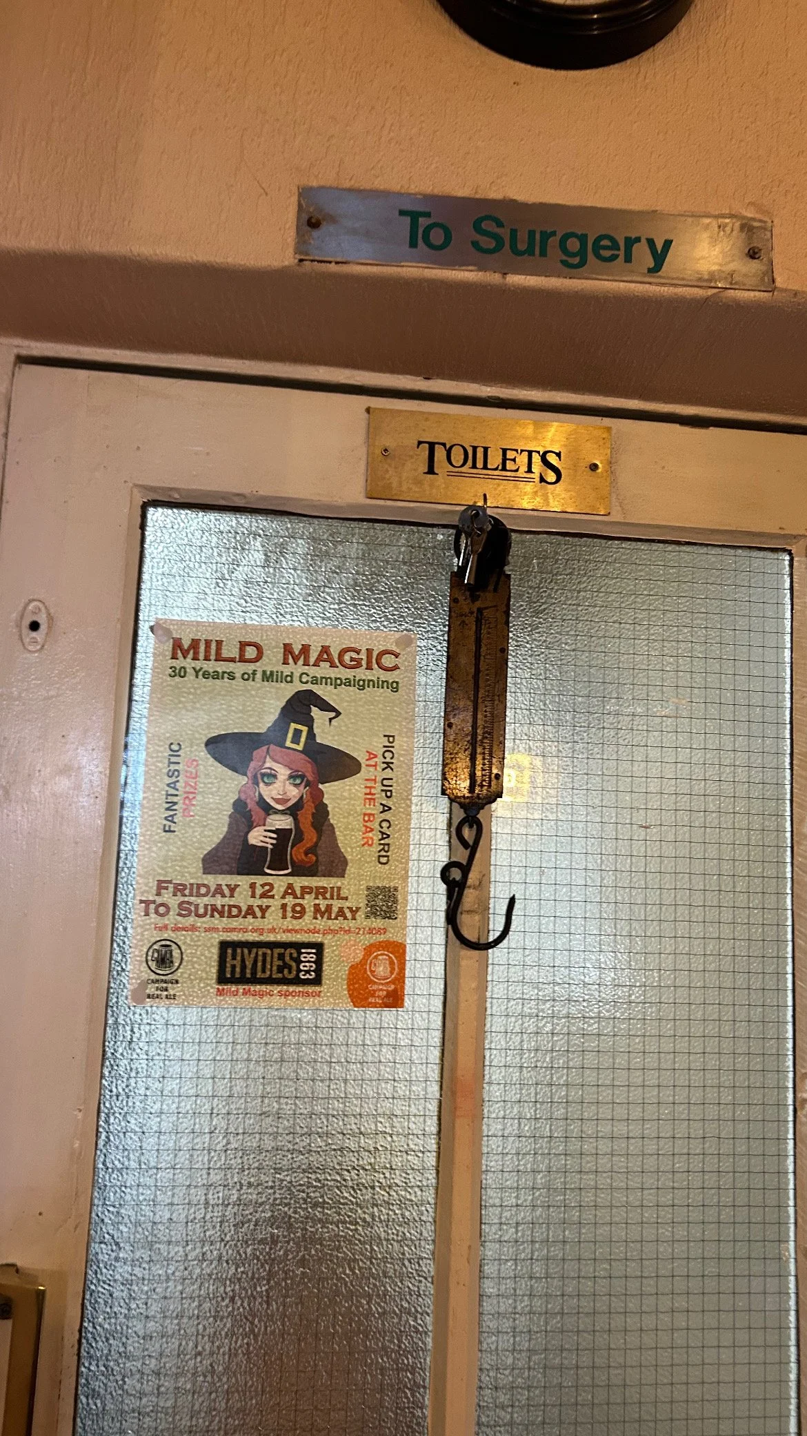 A door to the toilets with a glass panel, a poster advertising a magic-themed event, and a vintage hanging scale with a hook. Above the door is a sign pointing to surgery.