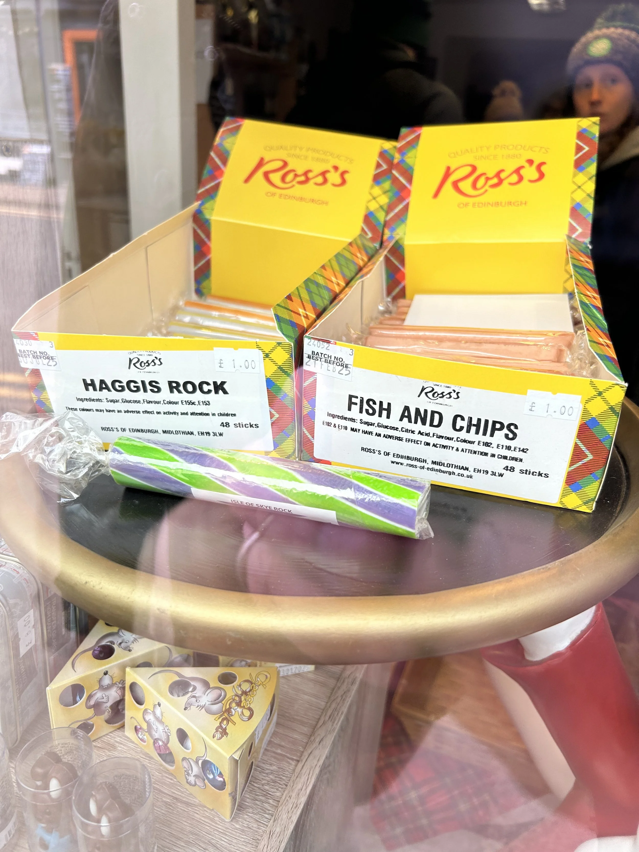 Two boxes of Scottish candy sticks labeled 'Haggis Rock' and 'Fish and Chips' on a display table, with a chocolate box and some small cups underneath. In the background, people are seen inside a store.