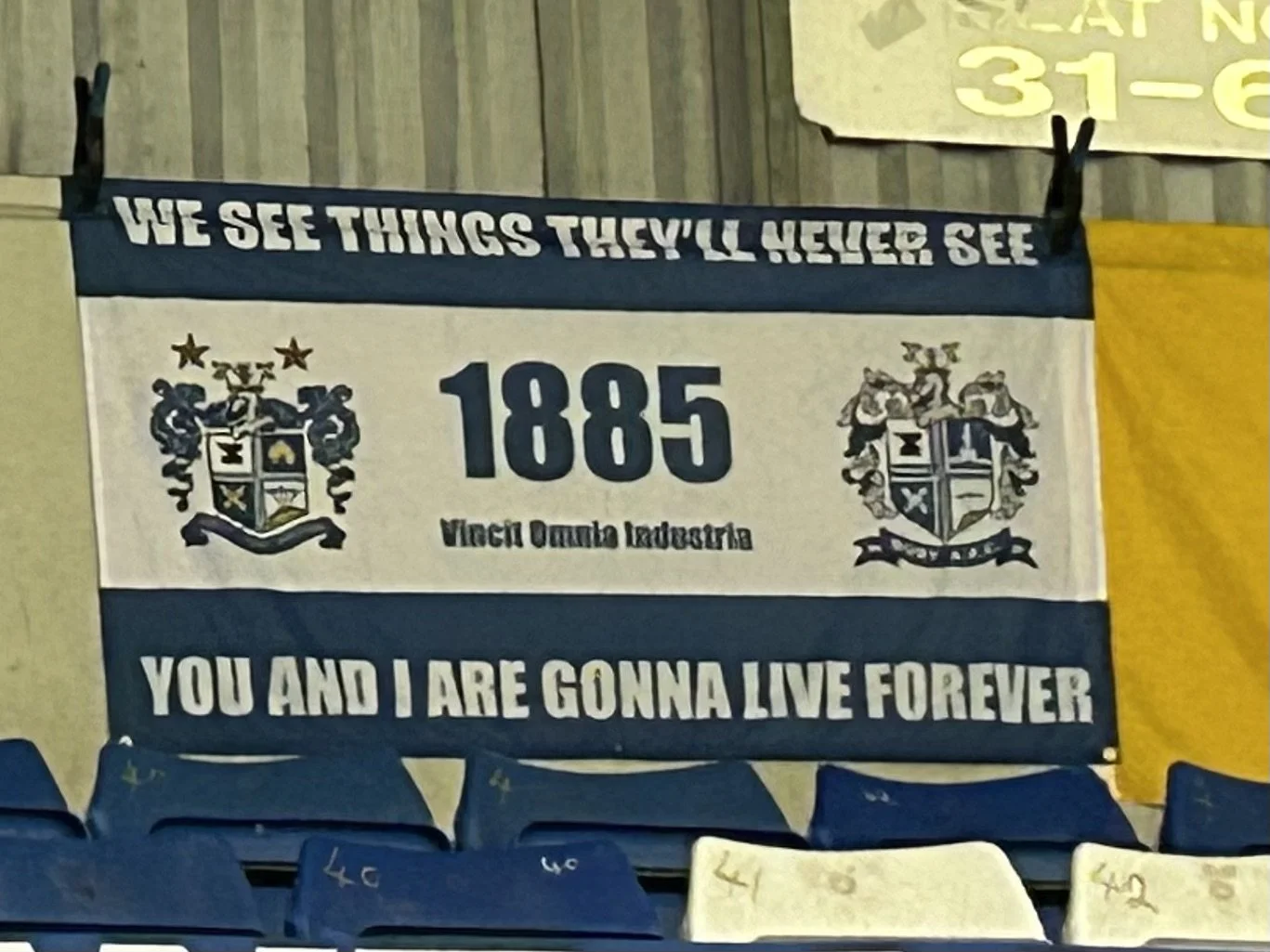 A banner with blue and white colors featuring a quote, coats of arms, and the year 1885, with the message "WE SEE THINGS THEY'LL NEVER SEE" and "YOU AND I ARE GONNA LIVE FOREVER".