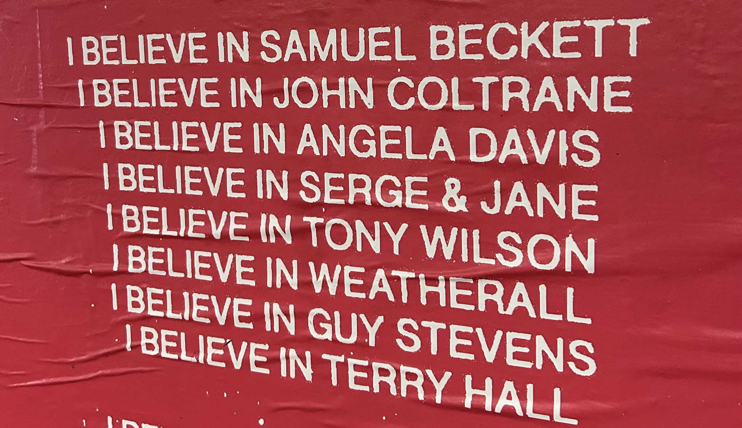 A red wall with white text listing names of individuals with the phrase 'I believe in' before each name.