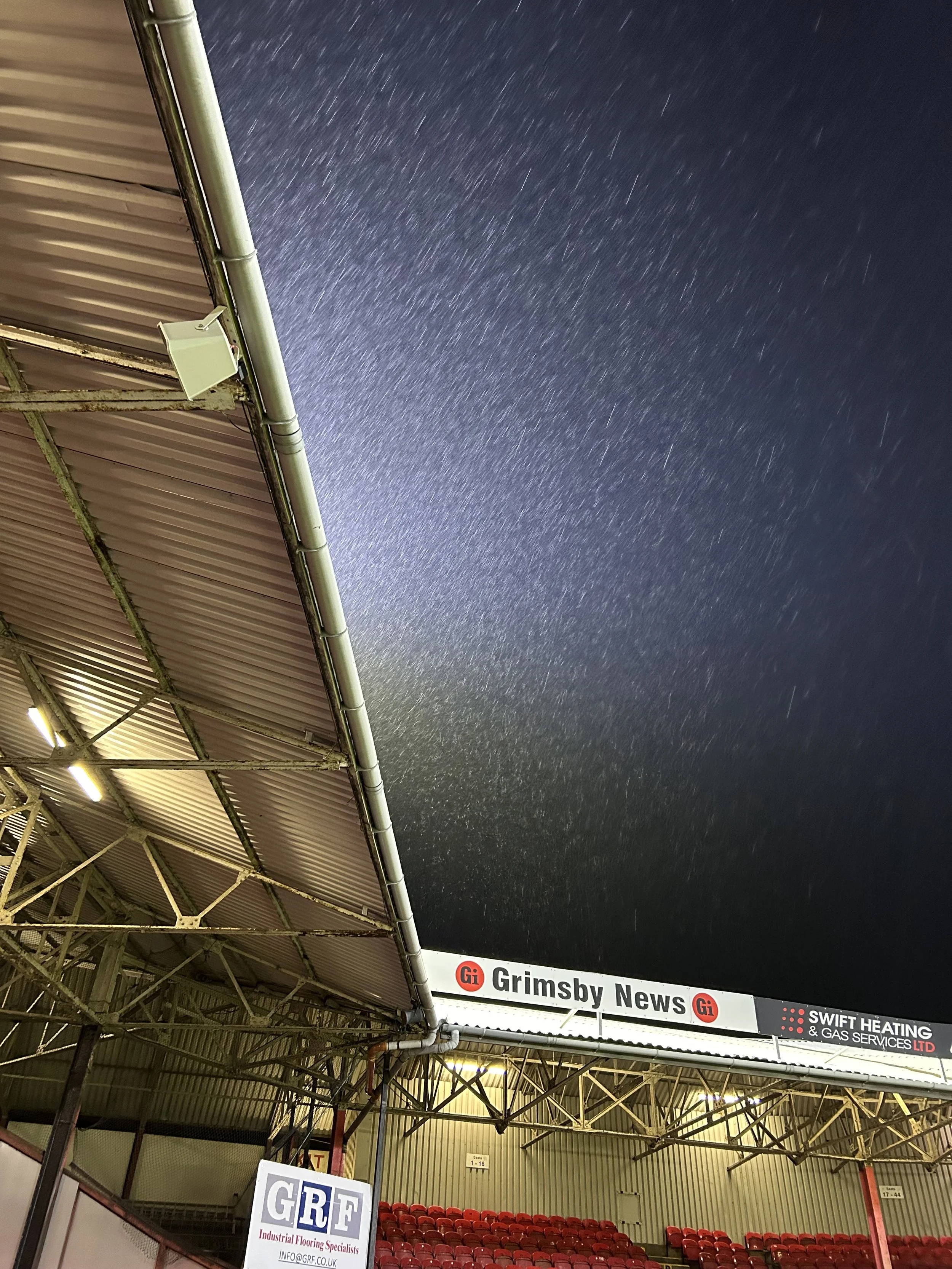 Night sky with star trails over a sports stadium or arena, visible seating and signage.