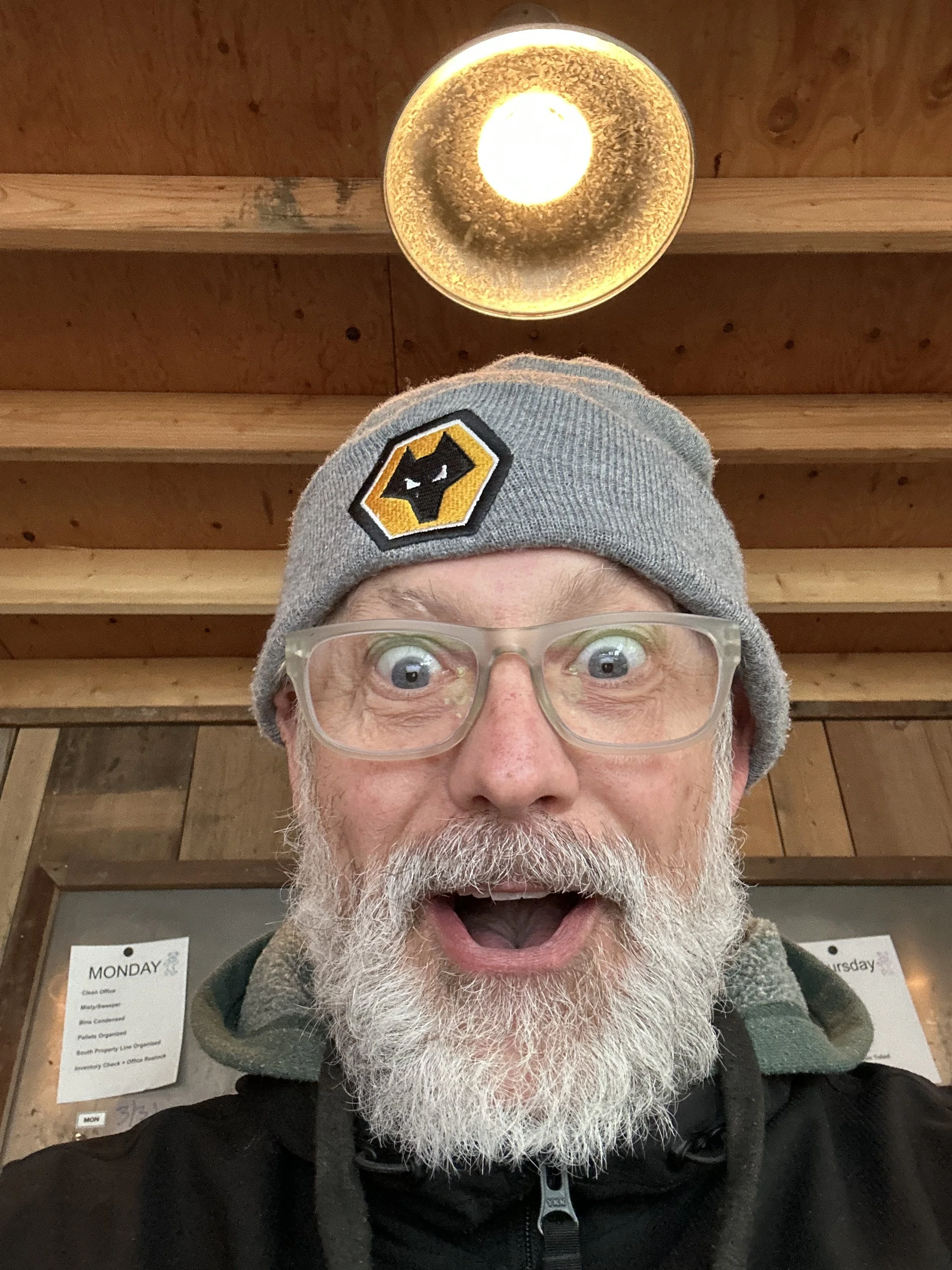 A man with glasses and a beard, wearing a gray beanie with a logo, making a surprised or excited facial expression in an indoor setting with wooden walls and ceiling, and a warm light fixture above his head.