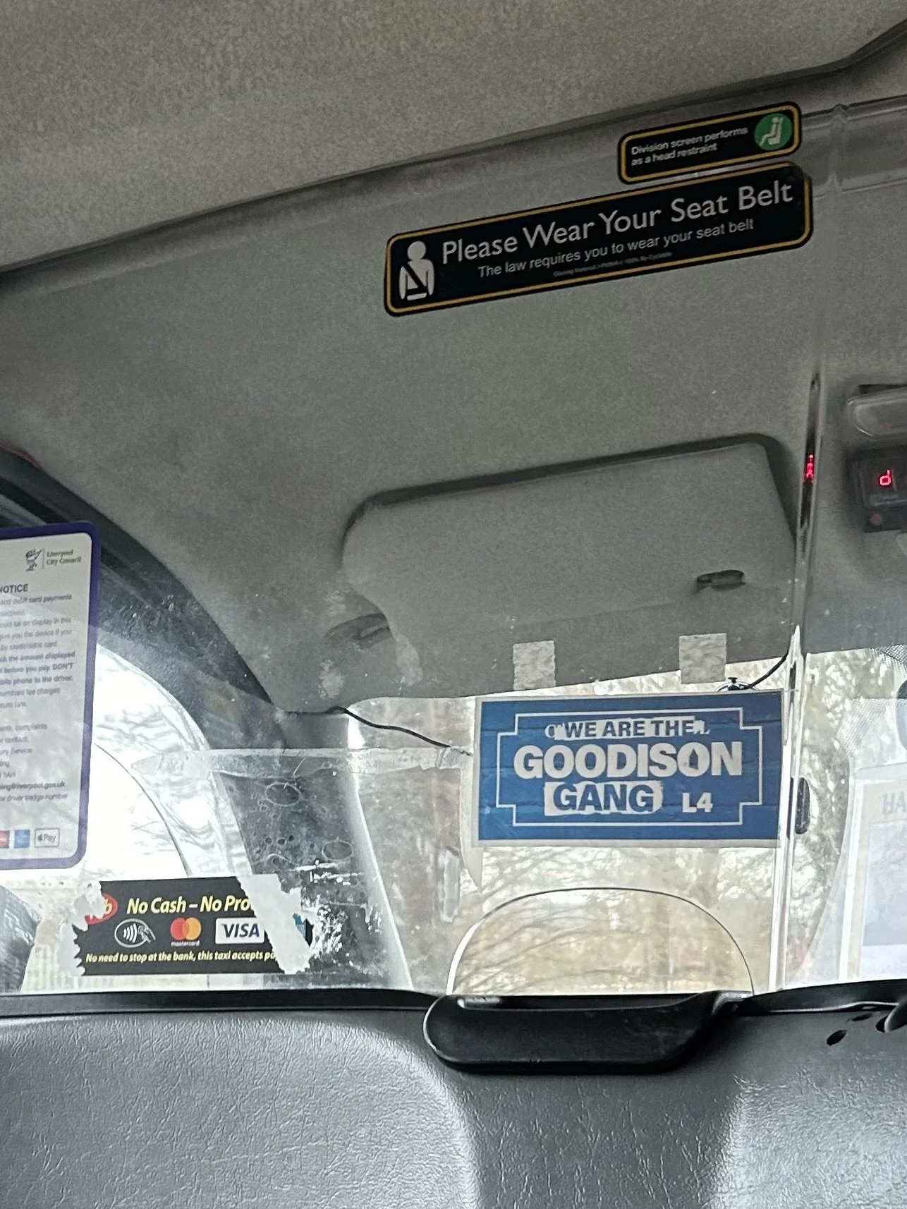 View from inside a taxi or bus showing a blue sign that says, 'WE ARE THE GOODISON GANG L4', and other signs including a seat belt reminder and payment options.