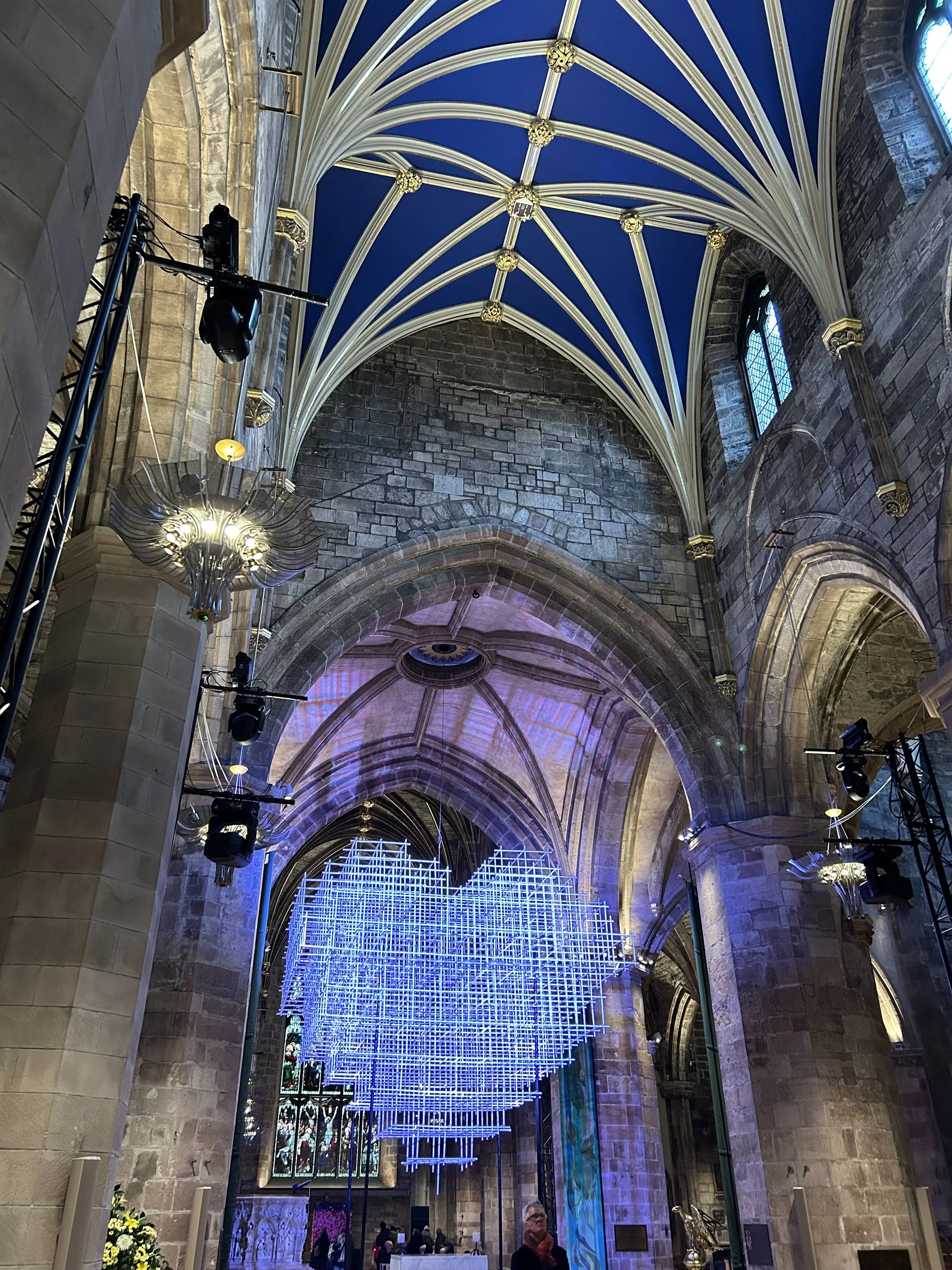 Interior of a historic church or cathedral with high vaulted ceilings, stone walls, stained glass windows, and modern light installation hanging from the ceiling.