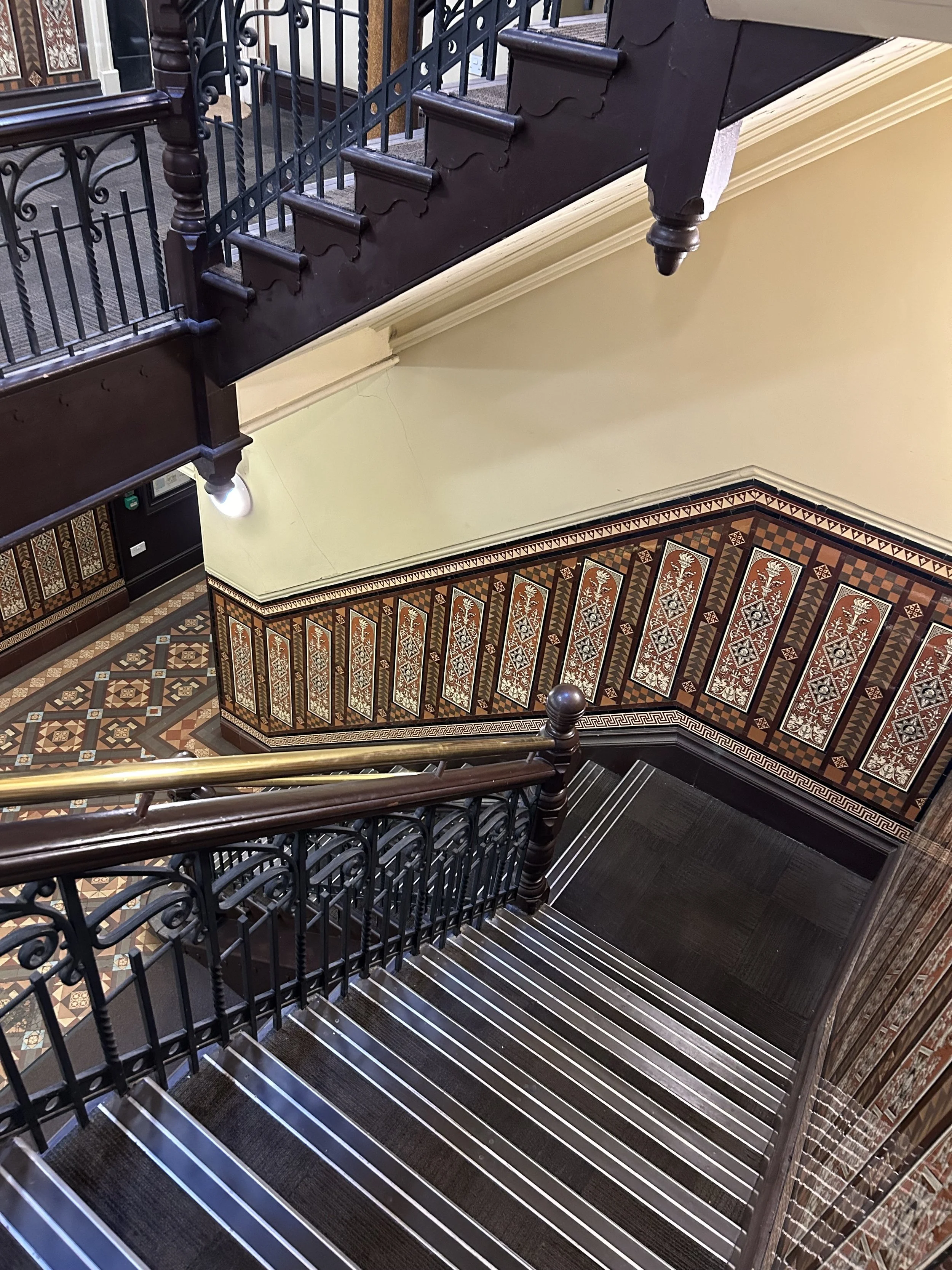 View of a vintage staircase with dark wood, metal railings, ornate patterned wall panels, and a patterned tiled floor.