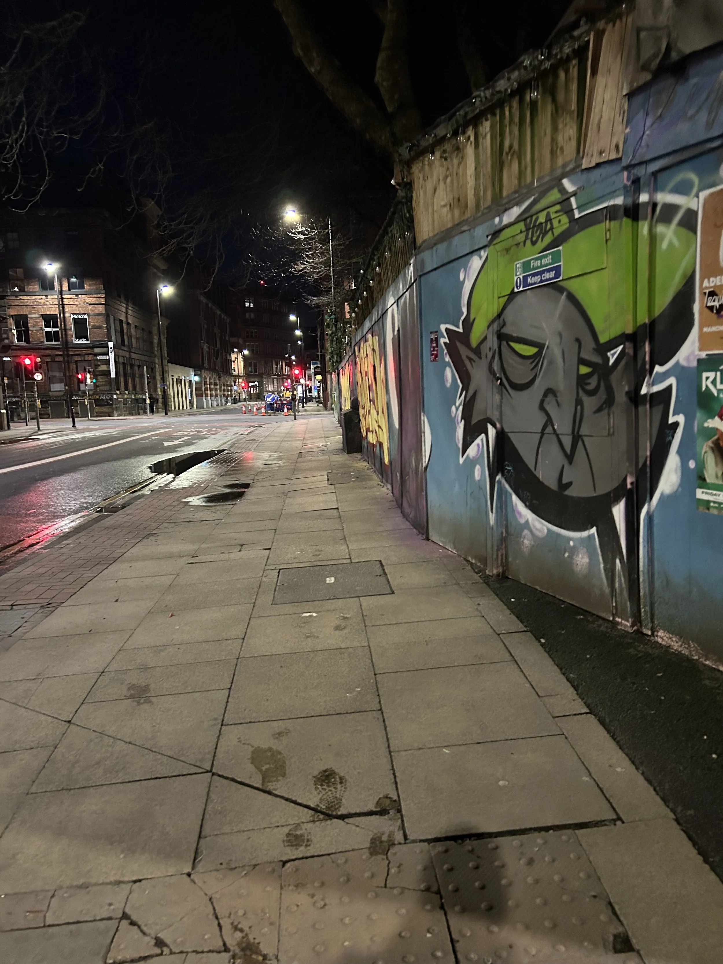 Empty city sidewalk at night with a colorful graffiti mural on a wall on the right side, wet pavement reflections, and streetlights in the distance.