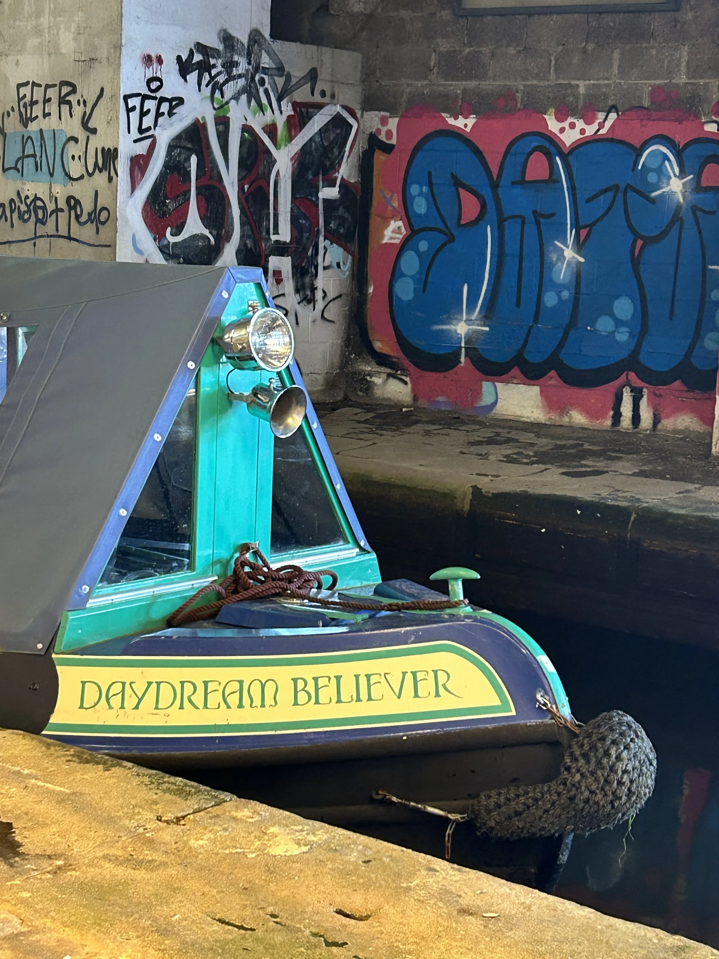 A small green and black boat labeled 'Daydream Believer' docked near a graffiti-covered wall, with ropes and a gray knitted item hanging off the boat.