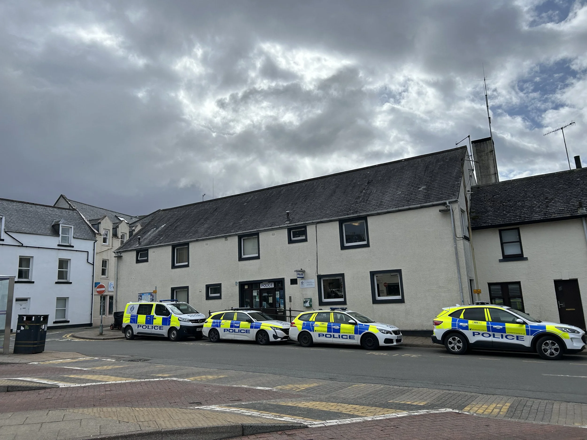 Four police vehicles parked in front of a white building on a street with cloudy sky overhead.