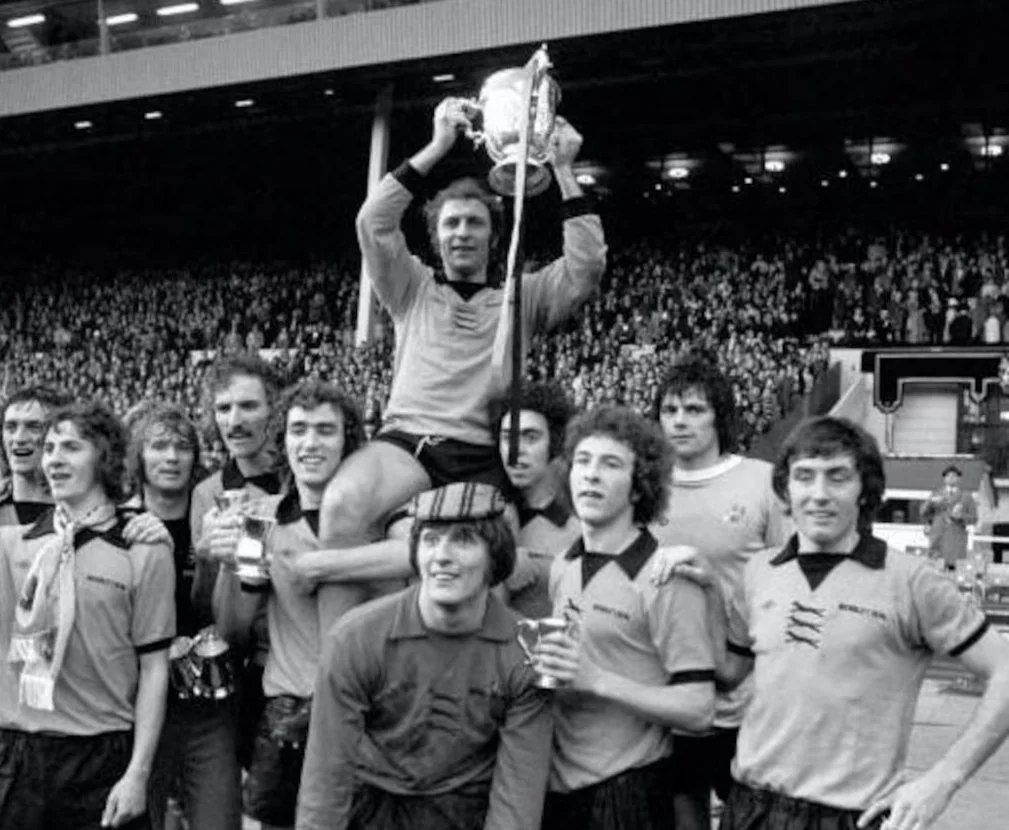 Men's soccer team celebrating victory after a match, with the goalkeeper holding a trophy in a stadium filled with spectators.
