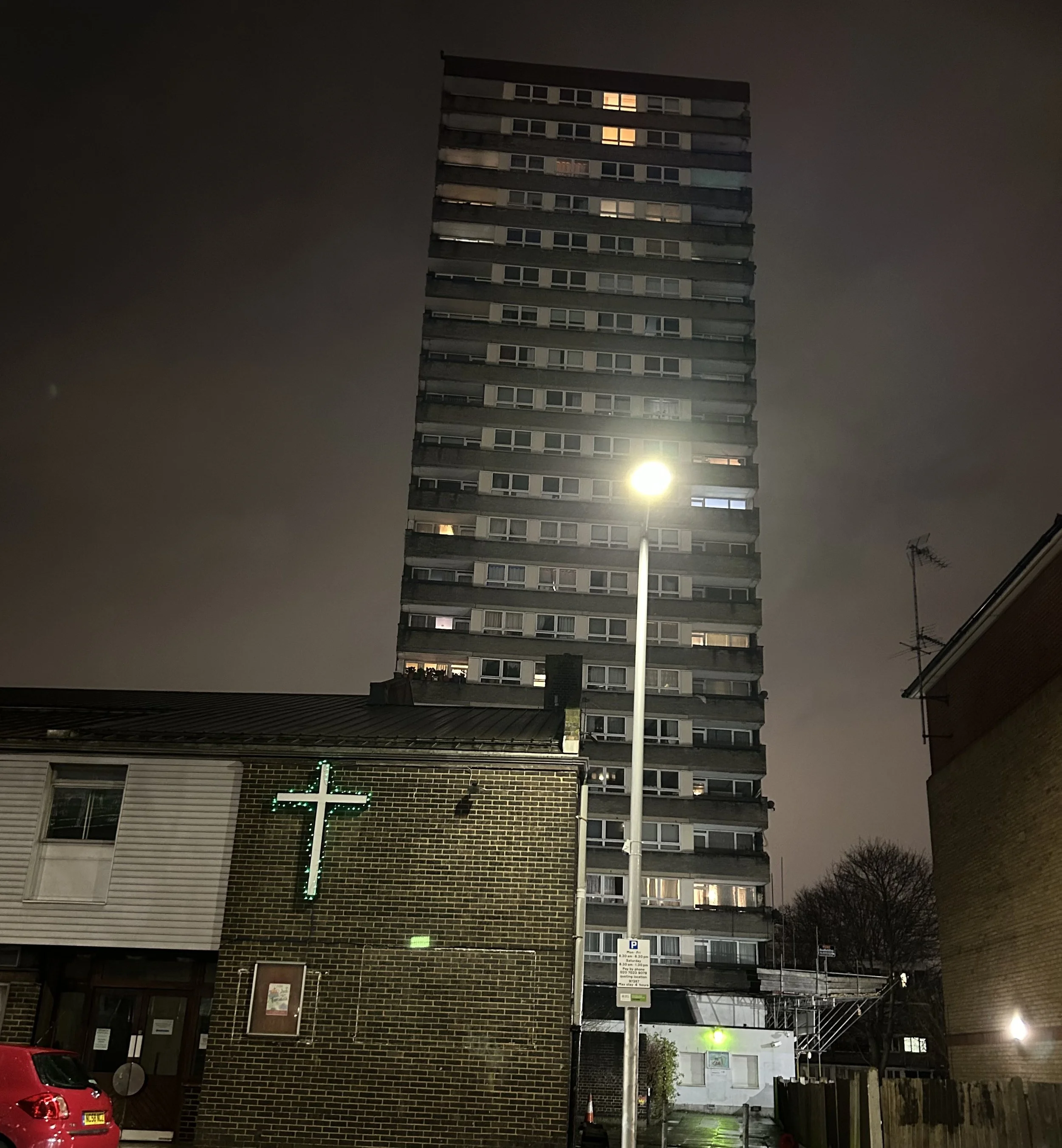 Nighttime city scene with a tall residential building, a church with a lit green cross, a streetlight, and parked cars. Cloudy sky in the background.